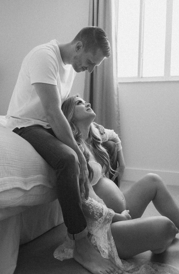 A romantic black and white boudoir photograph showing an intimate couple lounging together on a bed near a window.