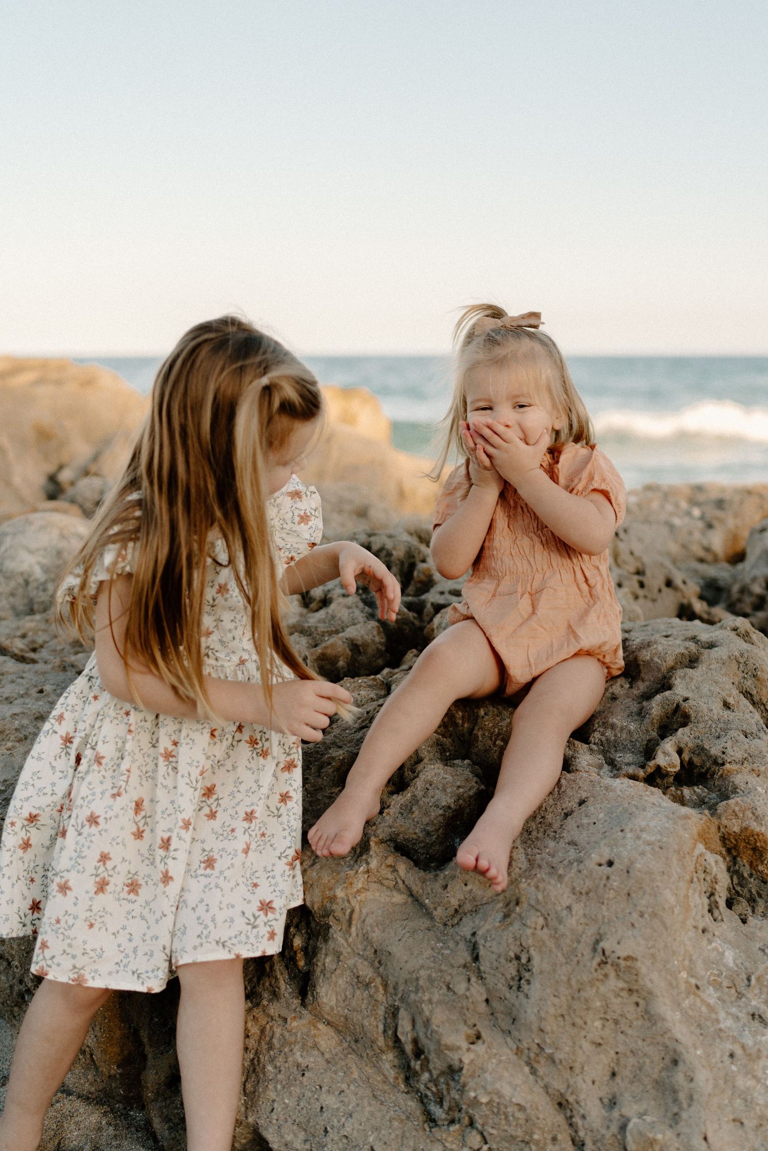 Two children in summer dresses share a playful moment on rocky coastal beach during sunset.