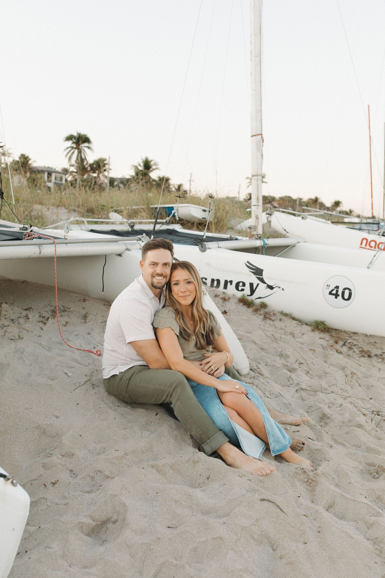 Smiling couple in casual clothes sits close together on a sandy beach in front of white sailboats with palm trees in the background.