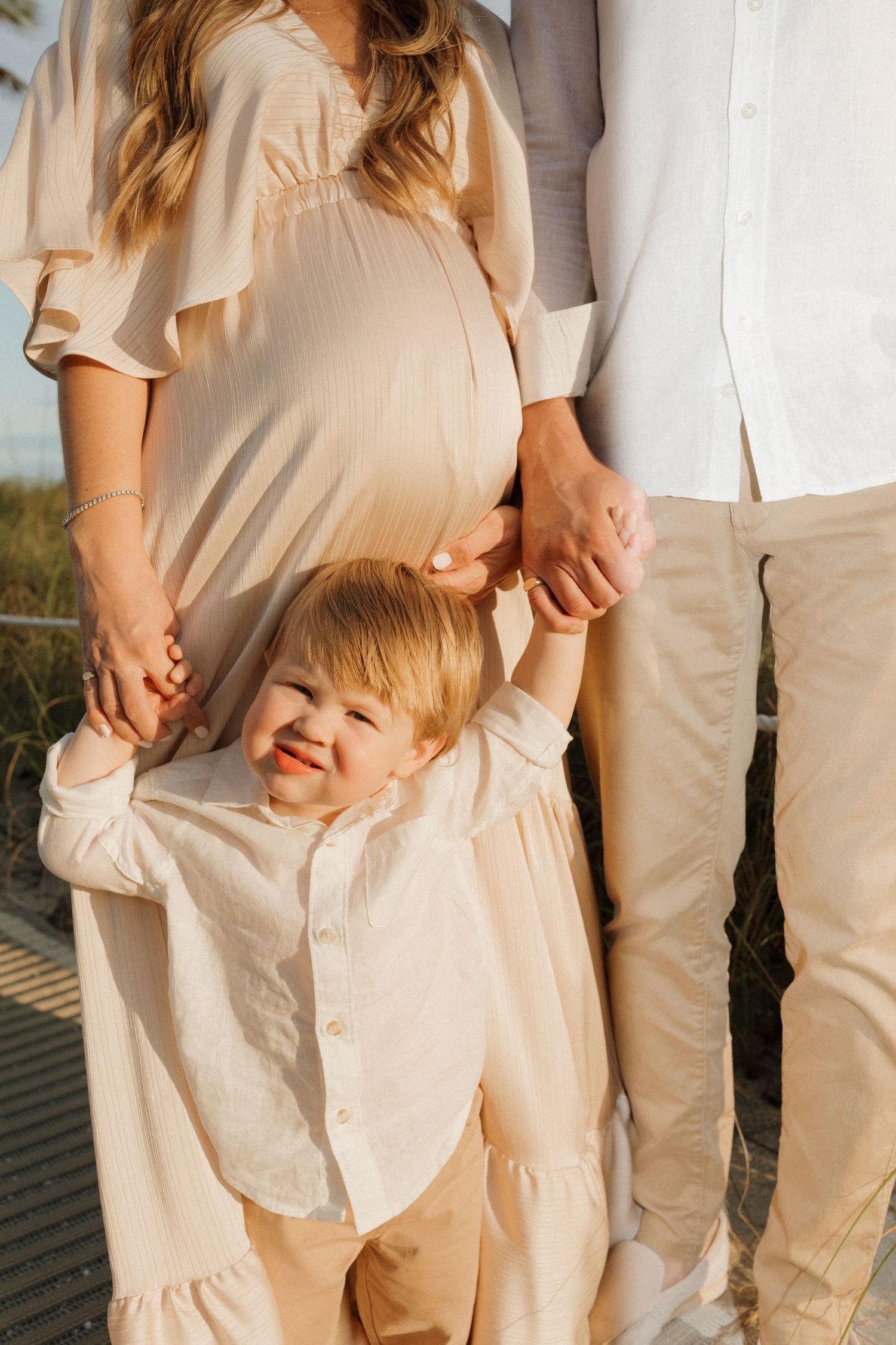 A joyful child in light clothing hugs their pregnant mother standing next to a person in white and beige outdoor.