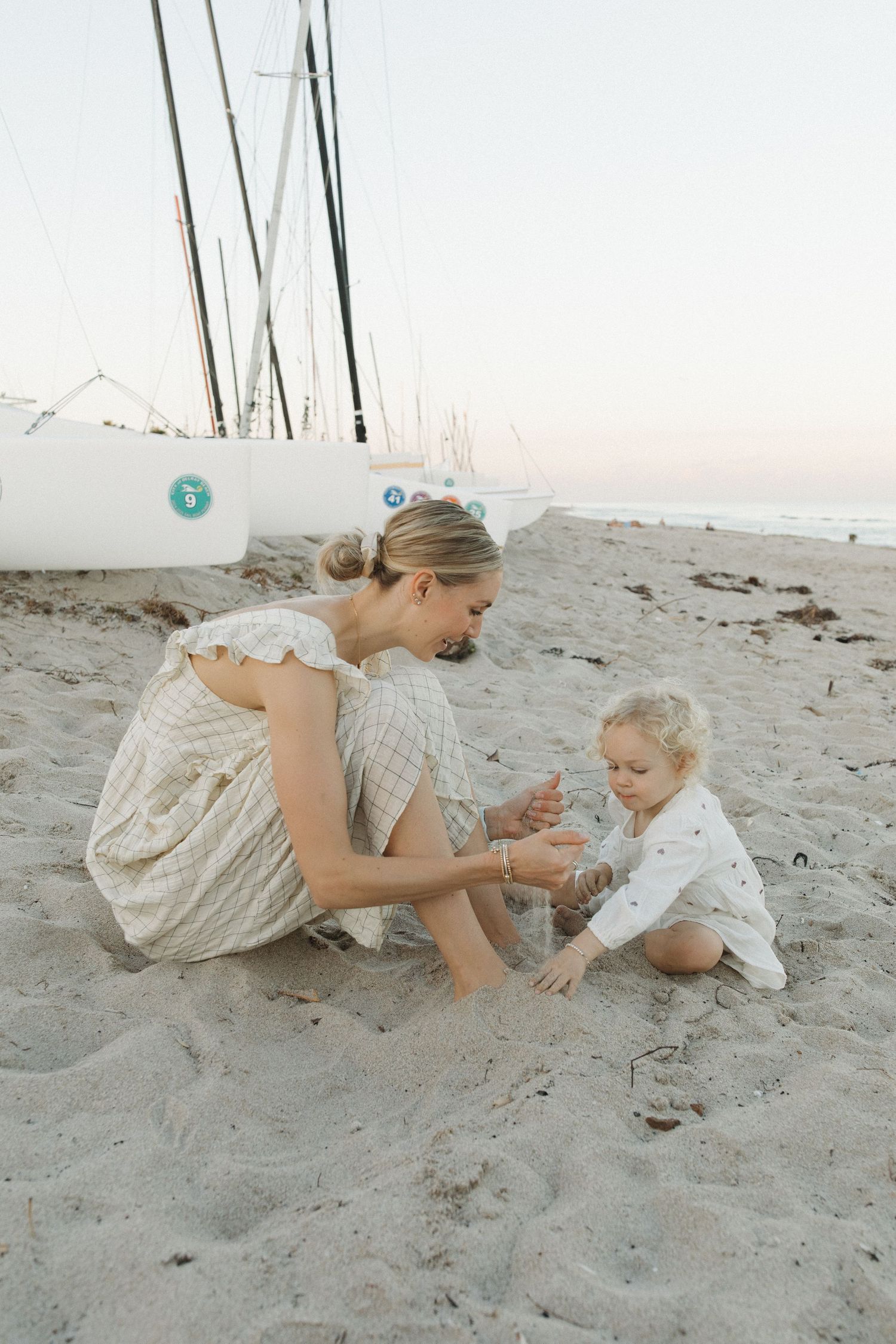 Woman in a flowy checkered dress and a toddler in white play together in the sand near sailboats on a beach at sunset.