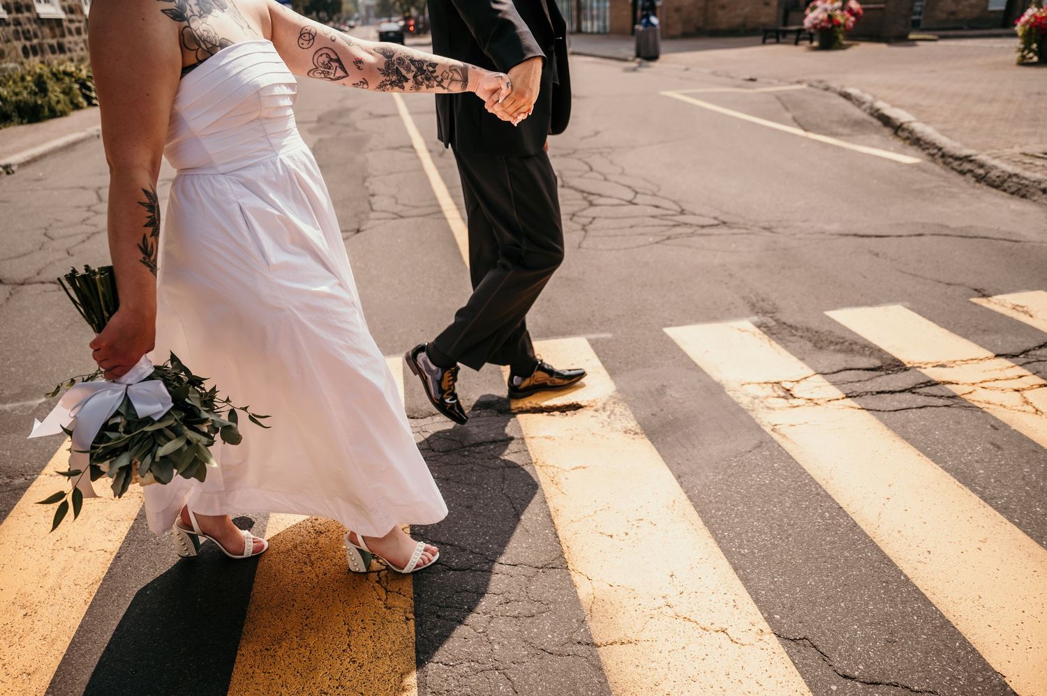 Couple walking across a crosswalk holding hands with the bride wearing a white wedding dress and holding flowers.