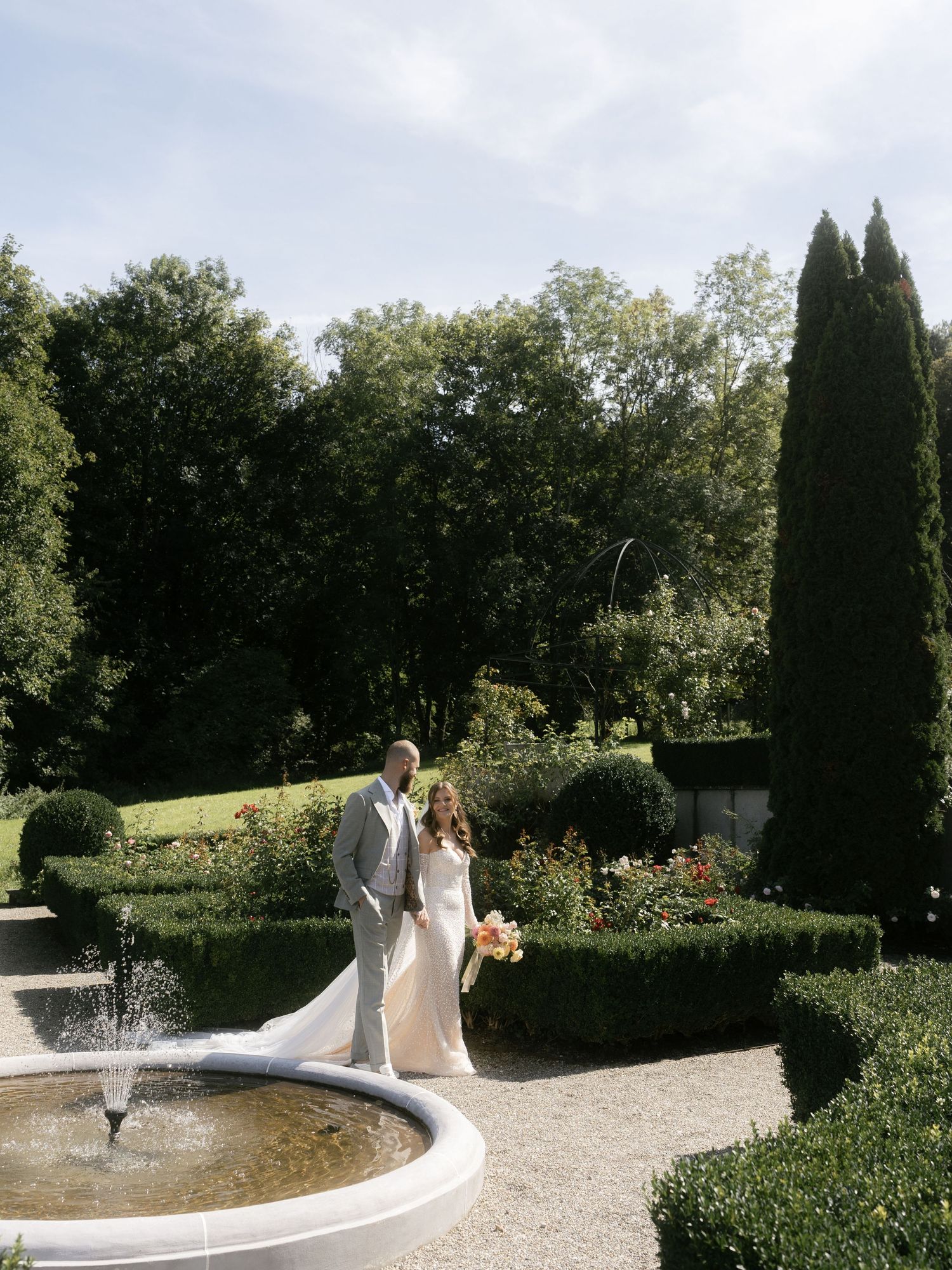 Couple in wedding attire walks along curved garden path with tall cypress trees and fountain in elegant outdoor setting.