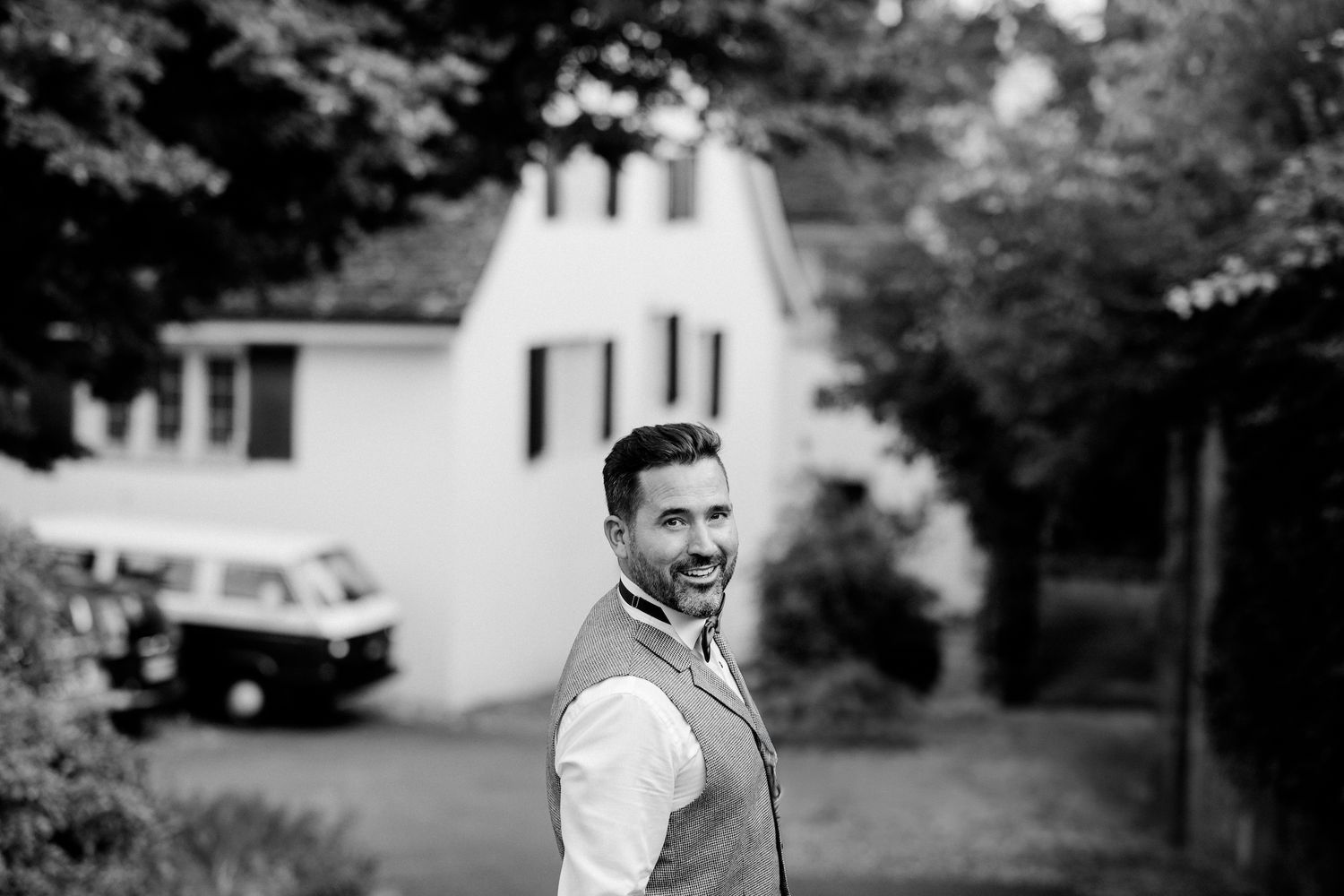 Smiling man in vest shirt standing outdoors in black and white photo with house and trees behind him.