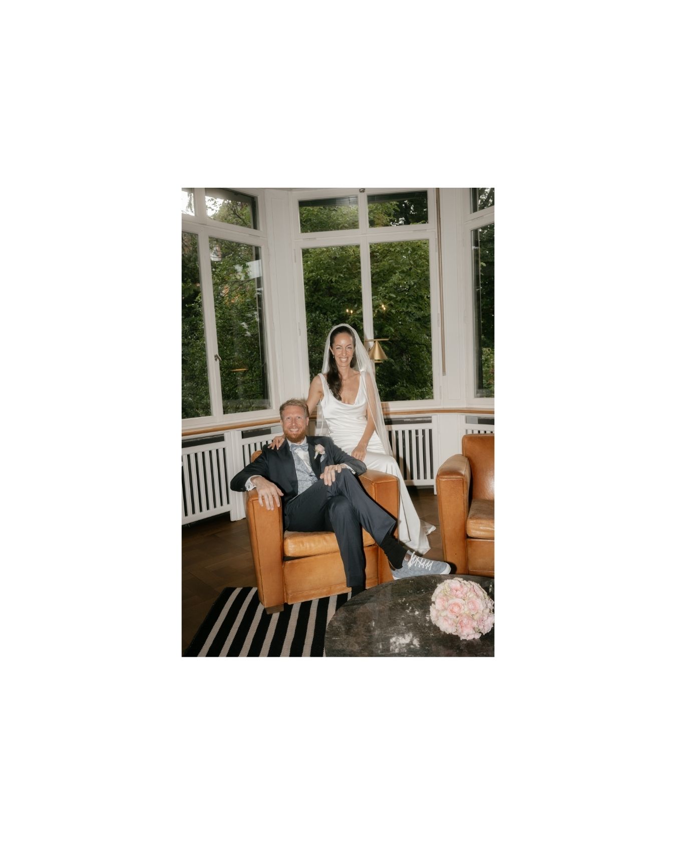 Bride standing behind groom seated in leather chair in sunlit room with striped rug and pink floral arrangement.
