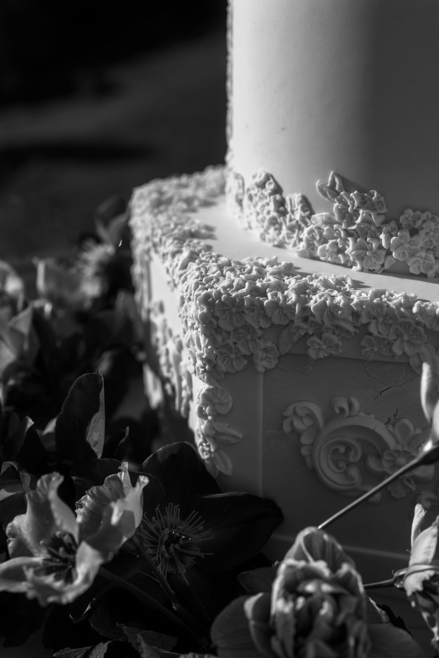 Black and white photo of an elegant decorated cake with ornate frosting details and a lit candle, surrounded by dark flowers.