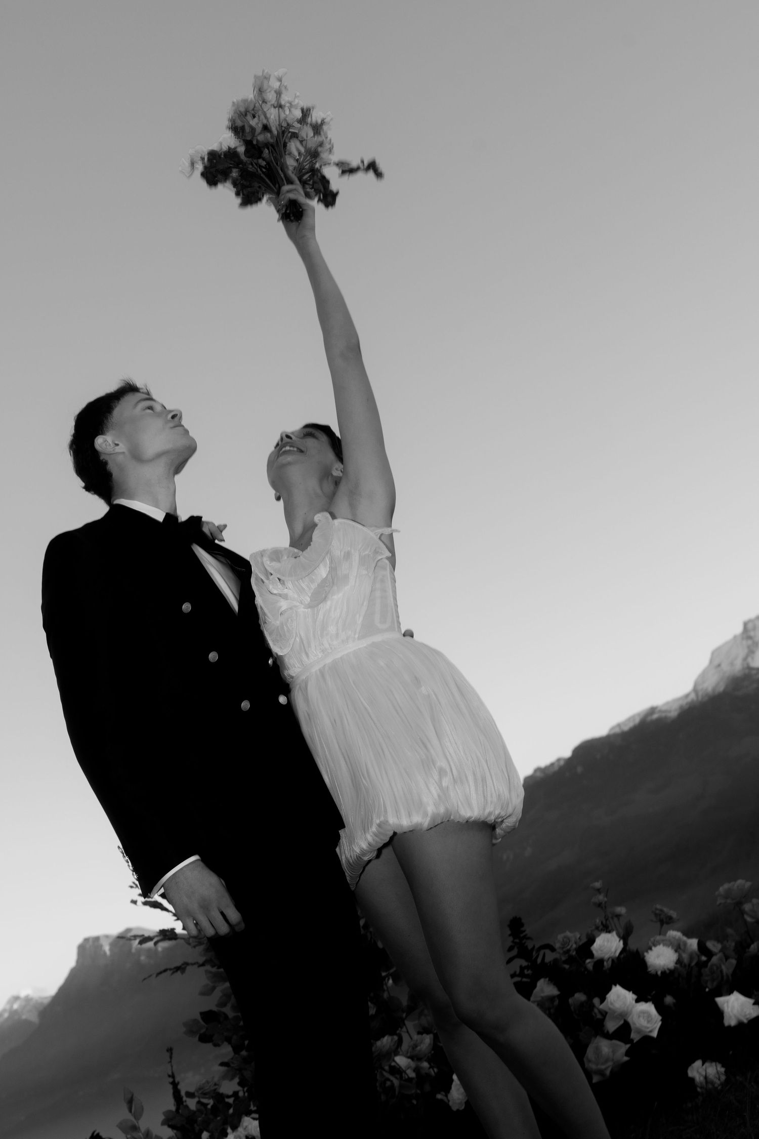 Black and white photo of a bride raising bouquet while groom looks up, mountains in background.