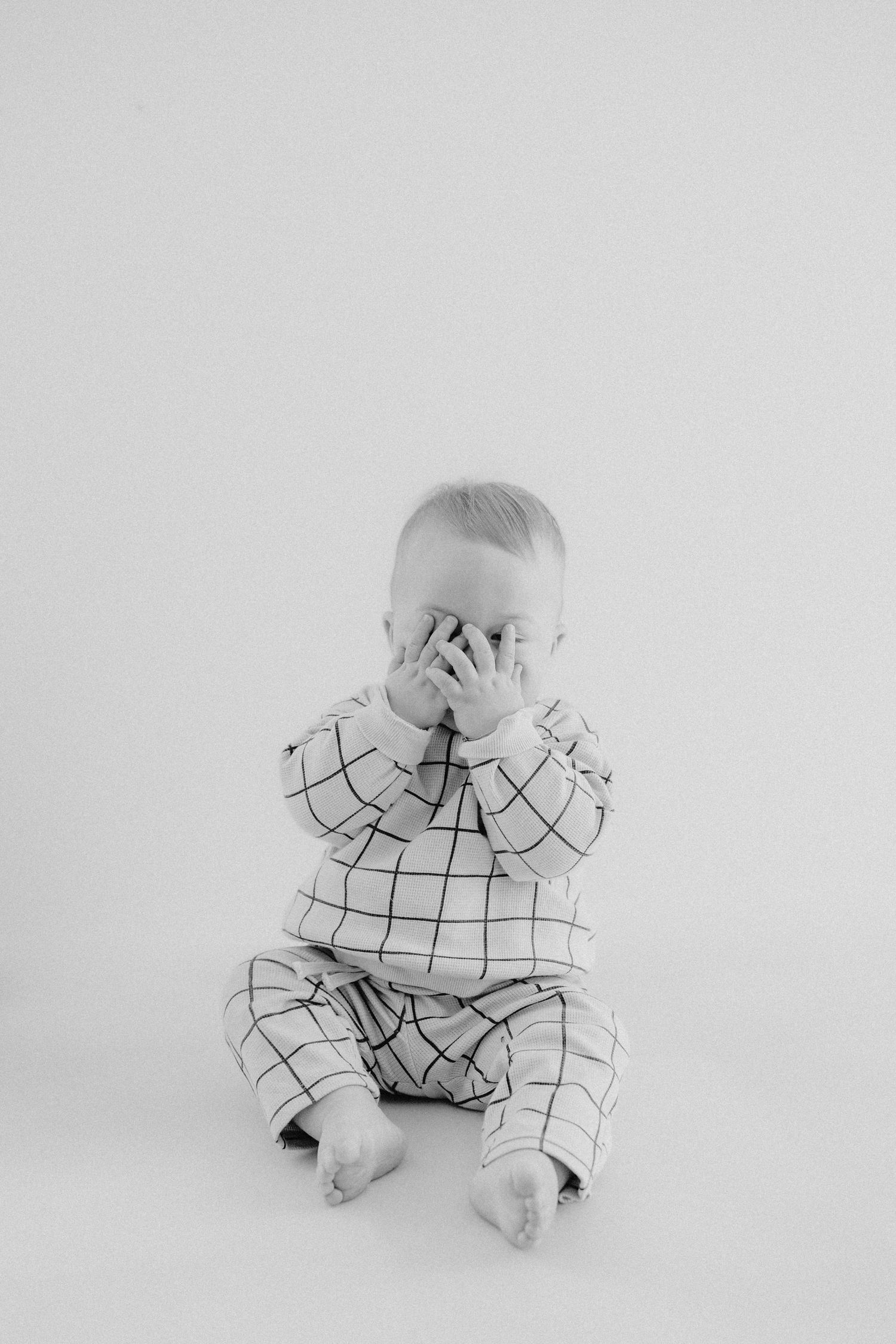 A small baby in a black and white checkered outfit sits on a white background in a minimalist photo series.