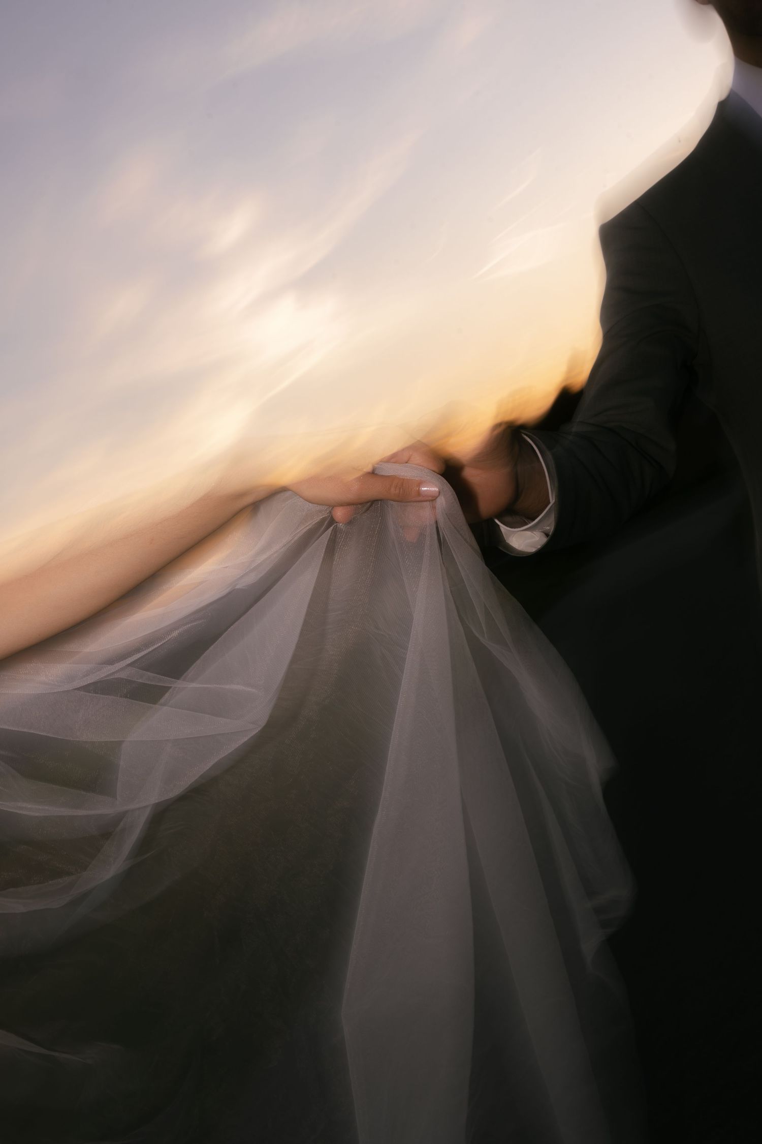 Bride and groom holding a flowing white veil together at sunset, captured with a dreamy motion blur effect.