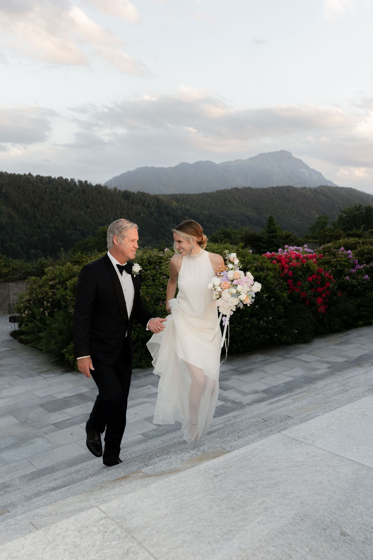 Bride in white gown holding floral bouquet walks with groom in tuxedo on terrace with mountain backdrop.