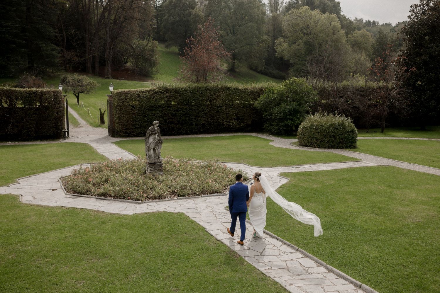 Bride and groom walk hand in hand through elegant formal garden with hedges and stone statue on their wedding day.