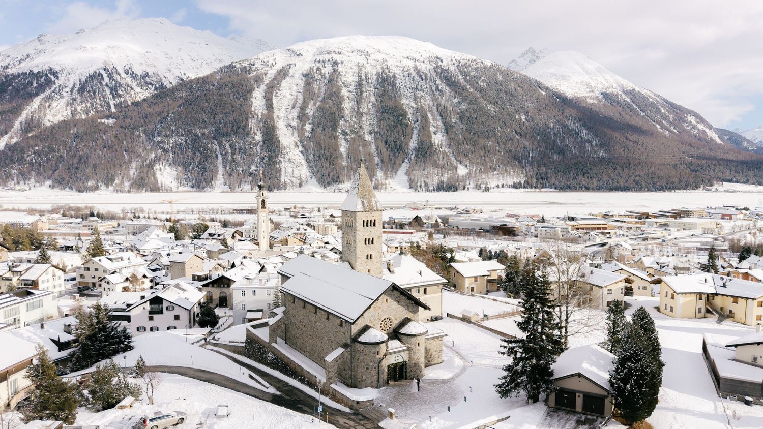 Snow-covered Alpine village with a church tower, surrounded by white mountains and frozen lake in winter.