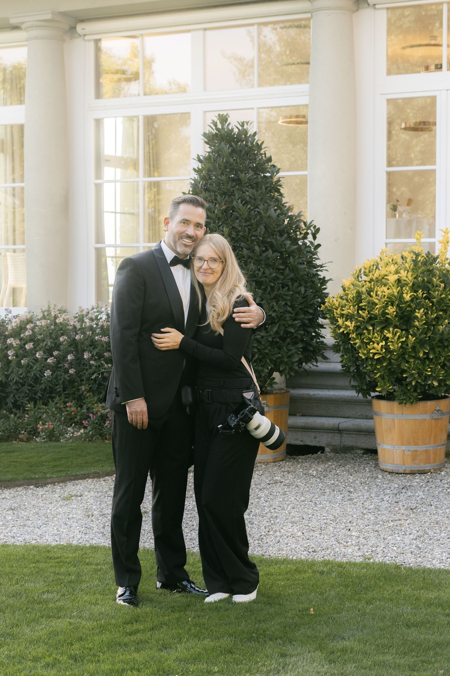 Couple in black formal attire embracing outdoors near elegant building with manicured hedges, woman holding camera.