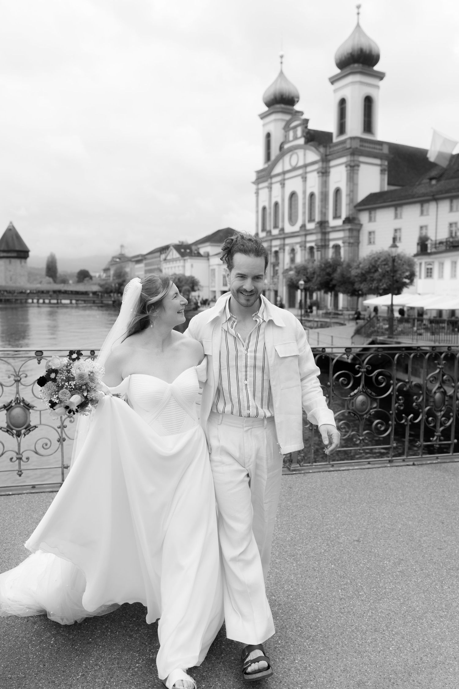 Black and white photo of a smiling bride and groom walking on a bridge in Lucerne, Switzerland, with Jesuit Church in background.