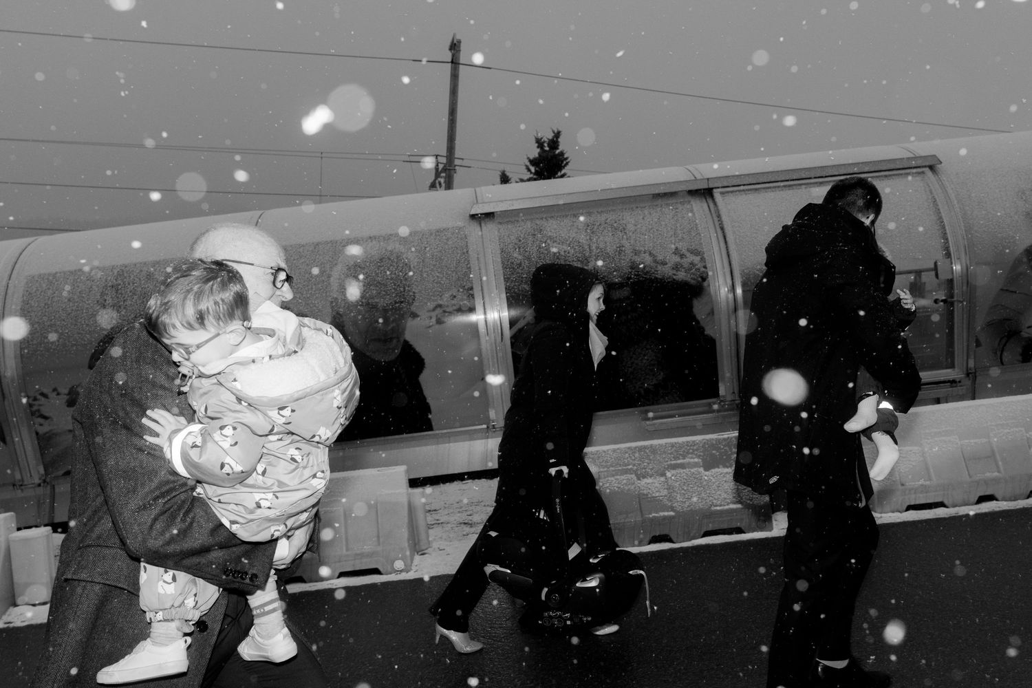Black and white photo of people carrying children in heavy snowfall near a bus during winter.
