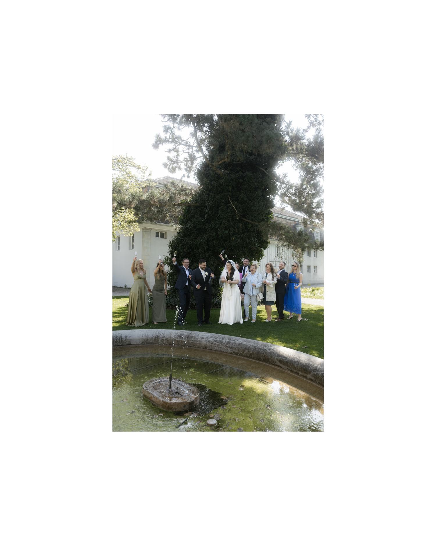 Wedding party posing near a decorative fountain in a lush garden with large trees and a white building.
