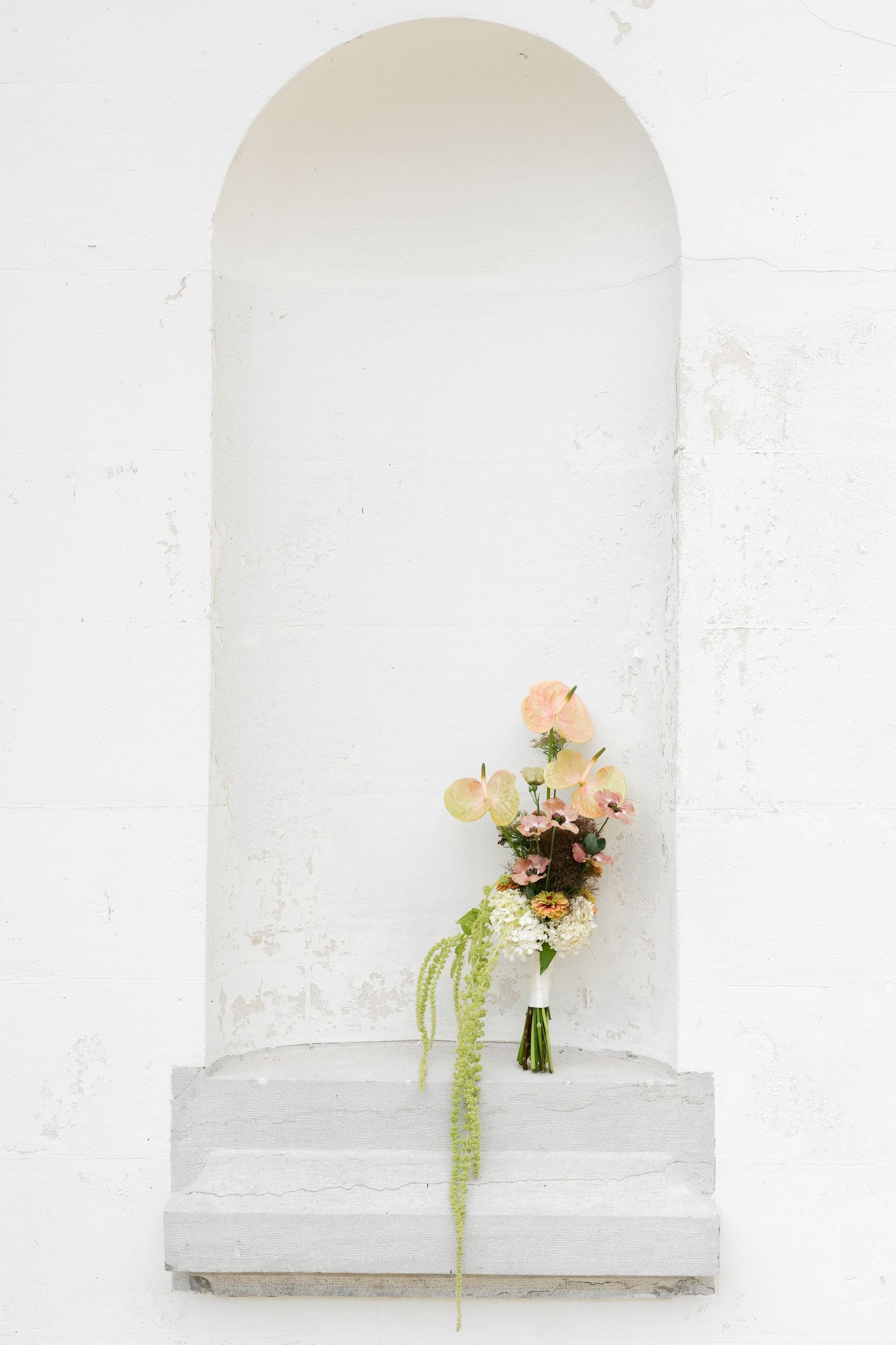 A delicate floral bouquet with pink roses and trailing greenery resting in a white arched niche on a textured wall.