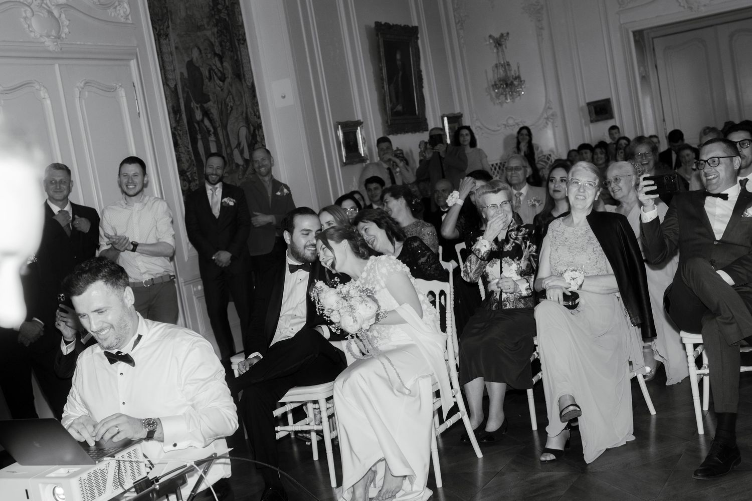 Black and white wedding reception photo showing guests seated and standing in an elegant ballroom.