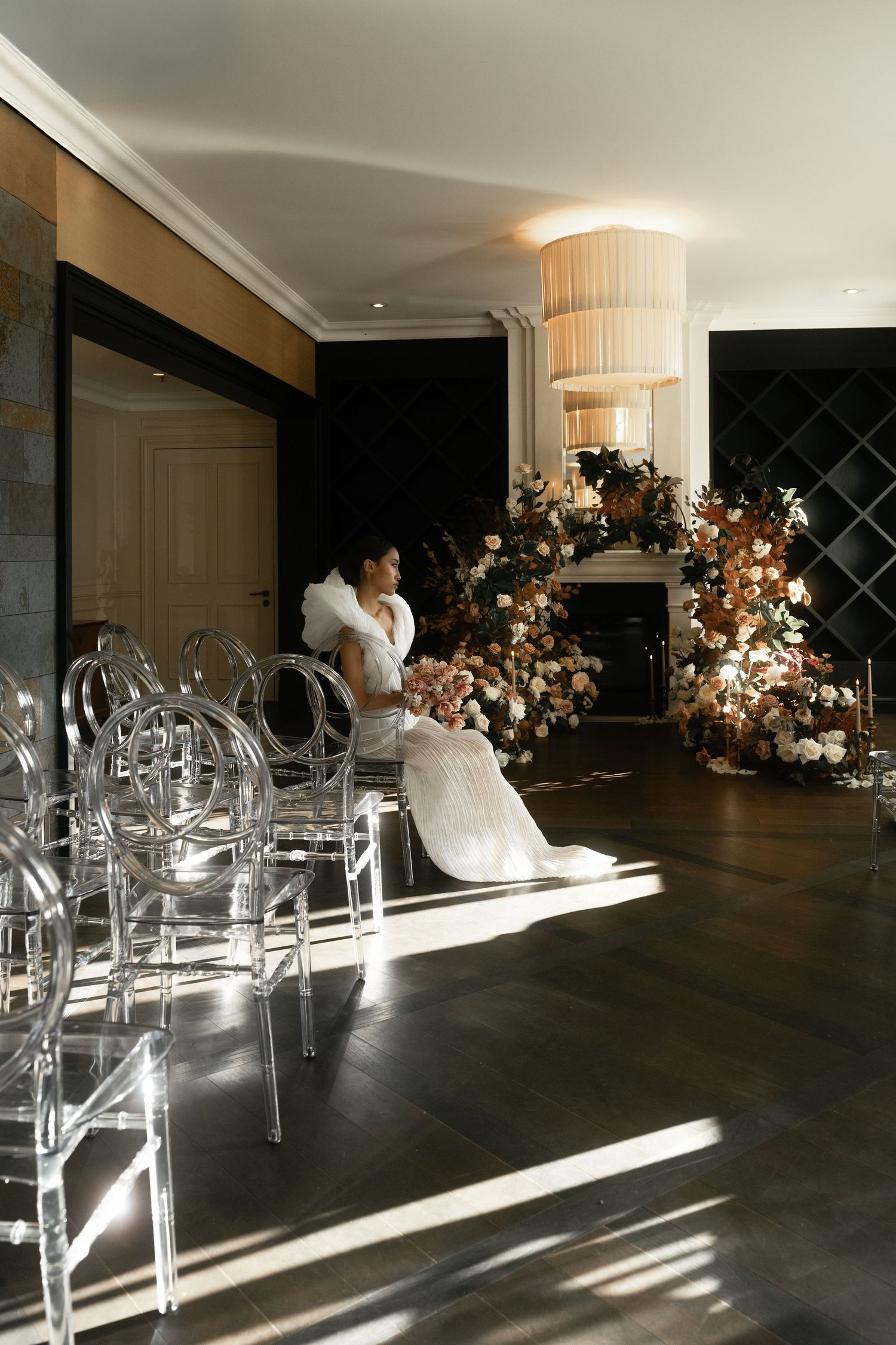 Modern wedding ceremony setup with clear acrylic chairs, elegant floral arrangements, and natural sunlight streaming across dark floors.