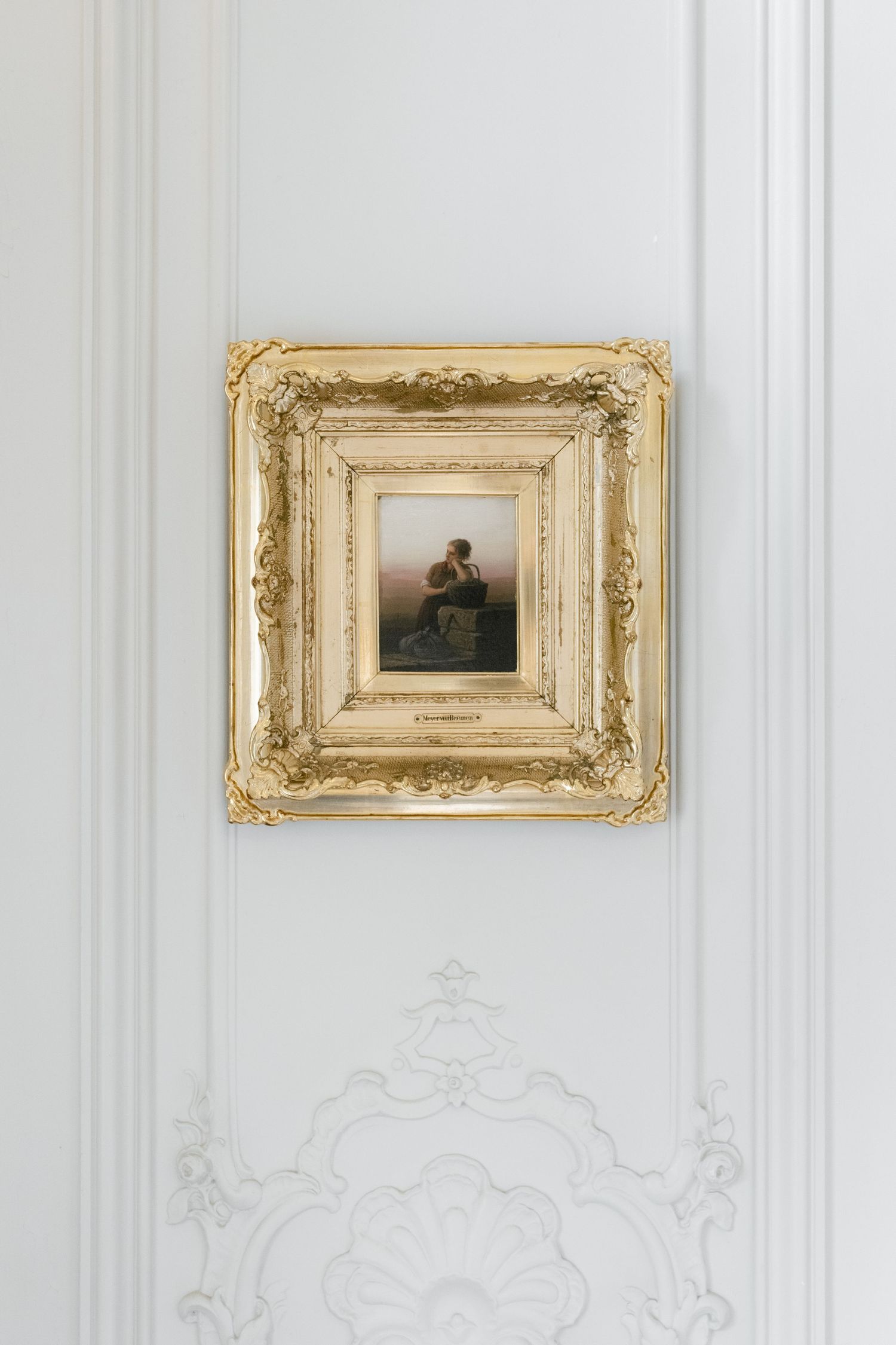 Small oil painting in ornate gold baroque frame mounted on white wall with decorative plaster molding below.