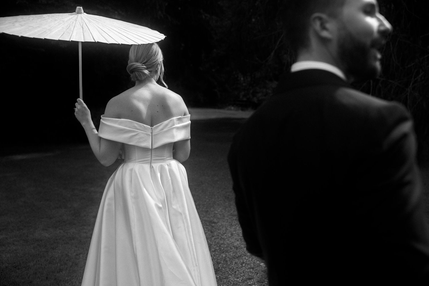Black and white wedding photo of bride in off-shoulder gown holding parasol, groom in tuxedo smiling in foreground.