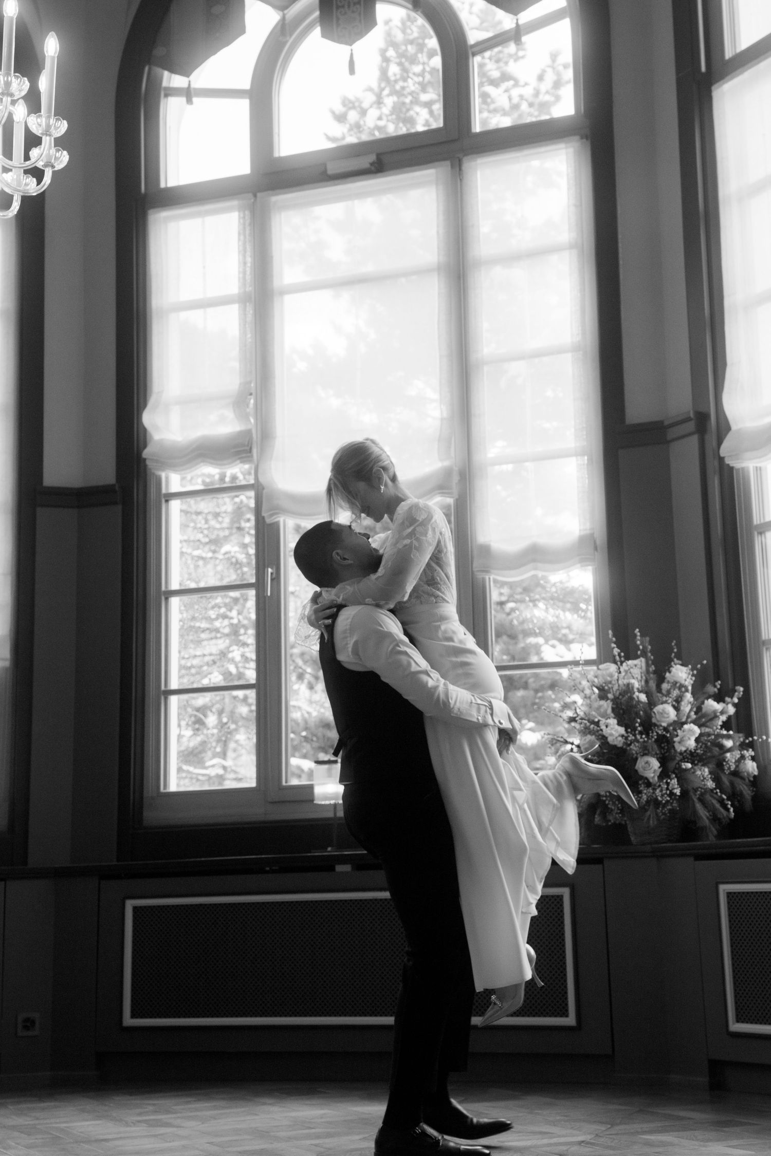 Black and white wedding photo of groom lifting bride in front of large arched window in elegant venue.