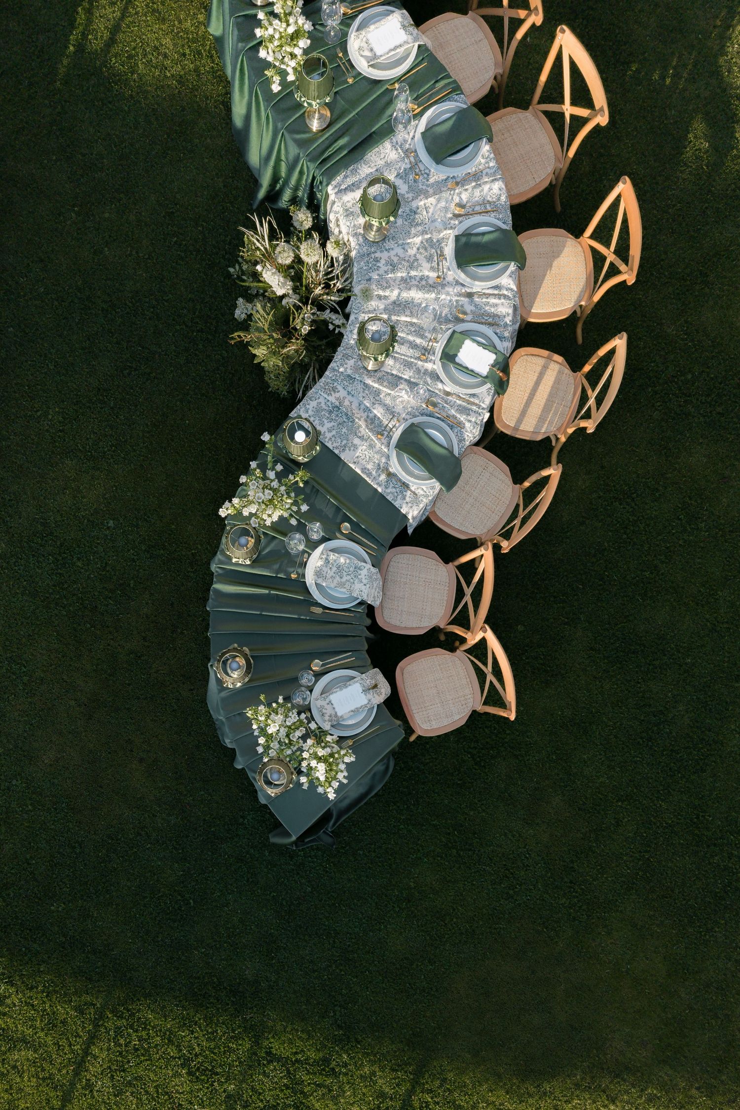 Aerial view of an elegant outdoor dining table setup with floral arrangements, dark linens, and wooden chairs on green grass.