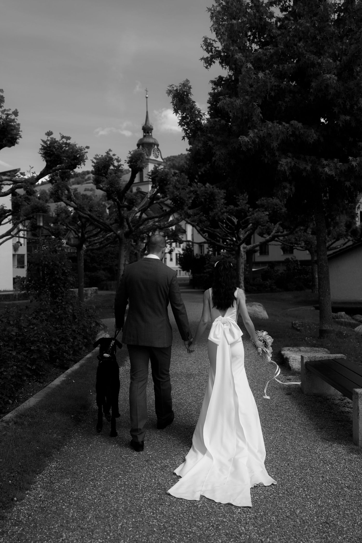 Black and white photo of bride and groom walking with dog toward church steeple, bride wearing gown with large bow.