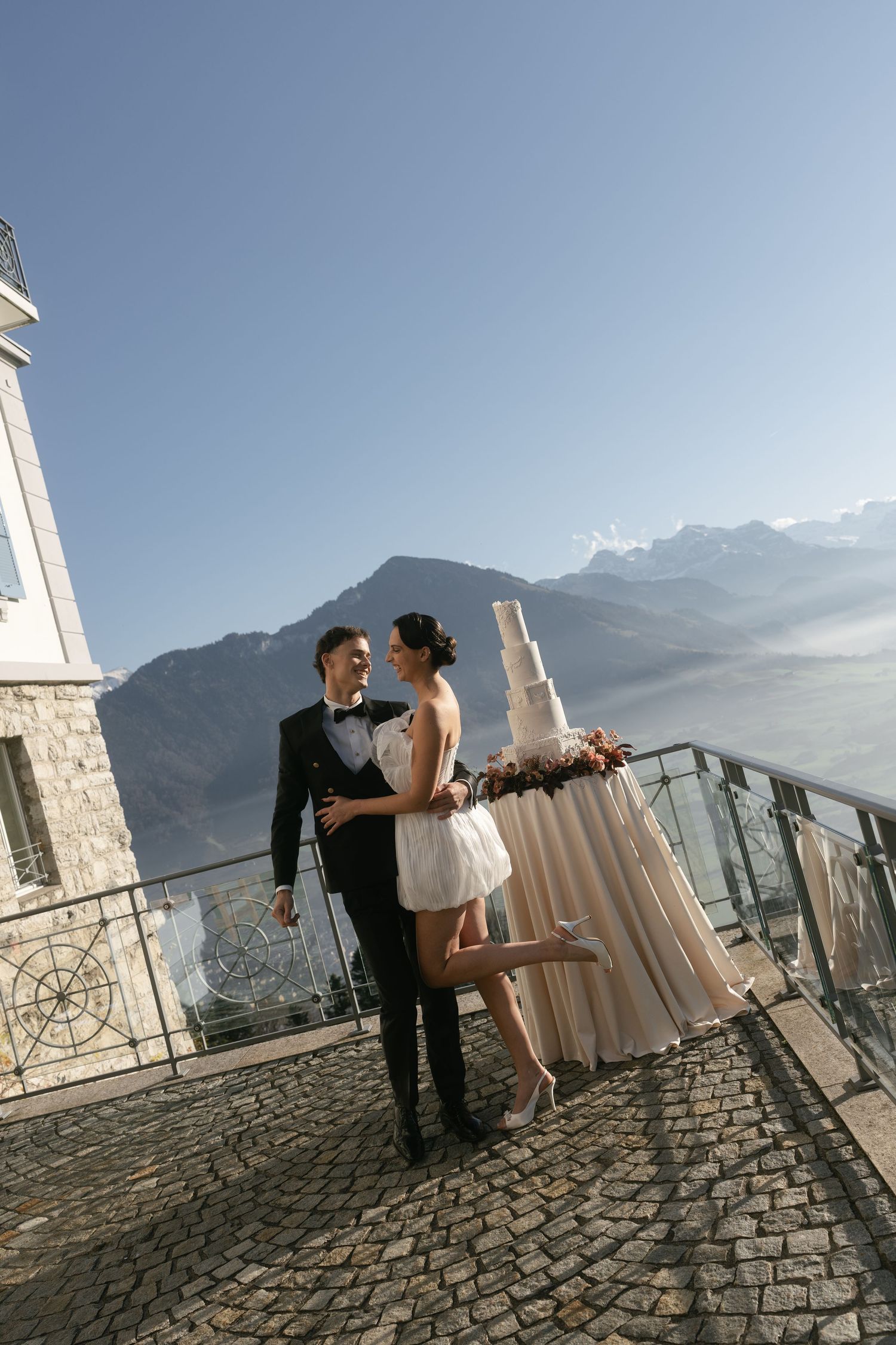 Bride and groom embrace on a scenic stone terrace with a tiered wedding cake, mountains and lake in the background.