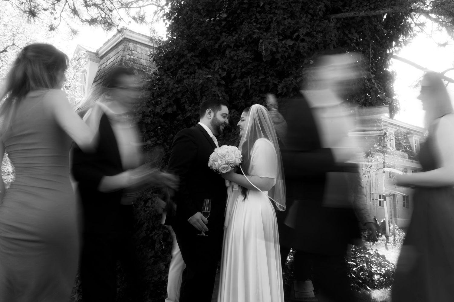 Black and white wedding photo of bride and groom kissing surrounded by blurred guests outdoors.