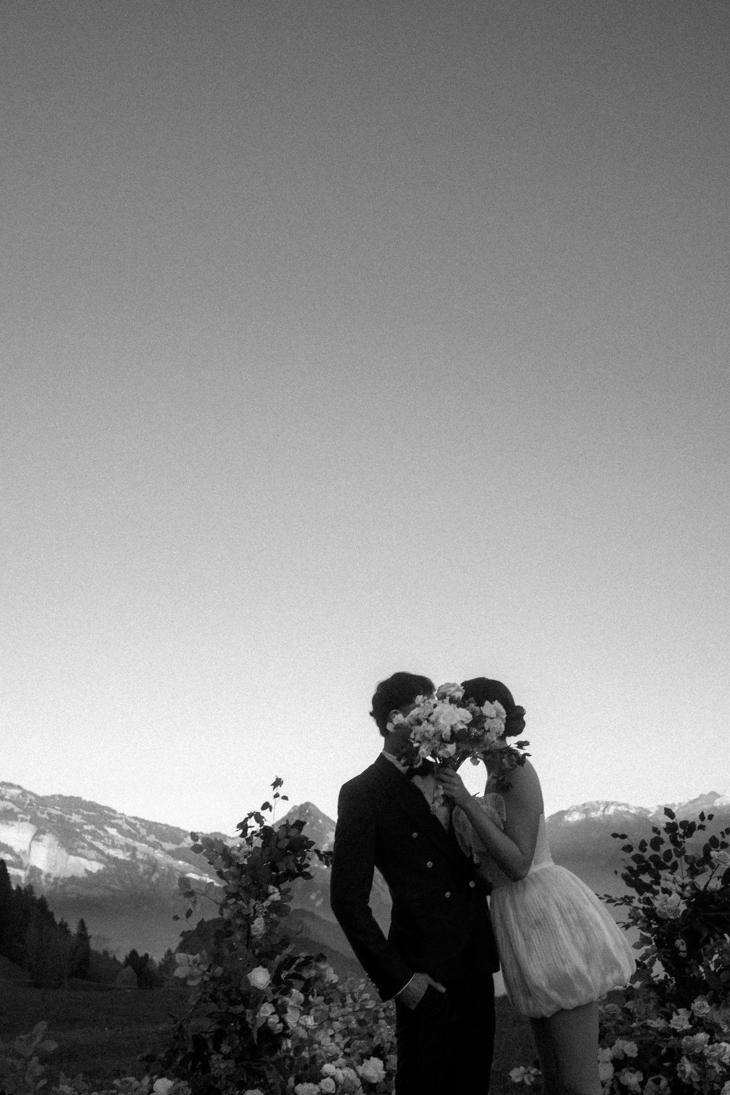 Black and white photo of a couple kissing behind a bouquet with mountain backdrop at their wedding at Villa Honegg