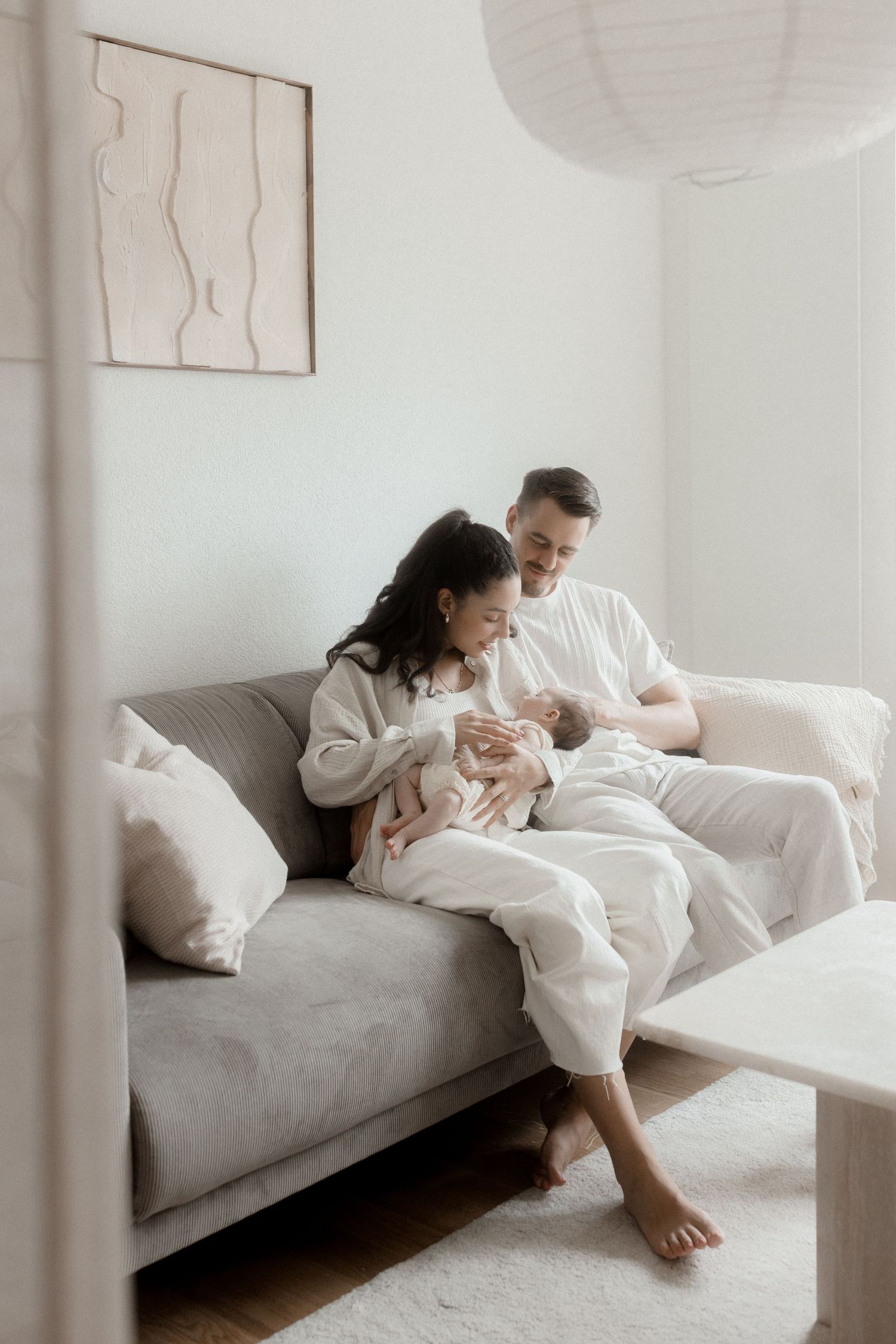 Family relaxing together on grey couch with newborn in minimalist white room.