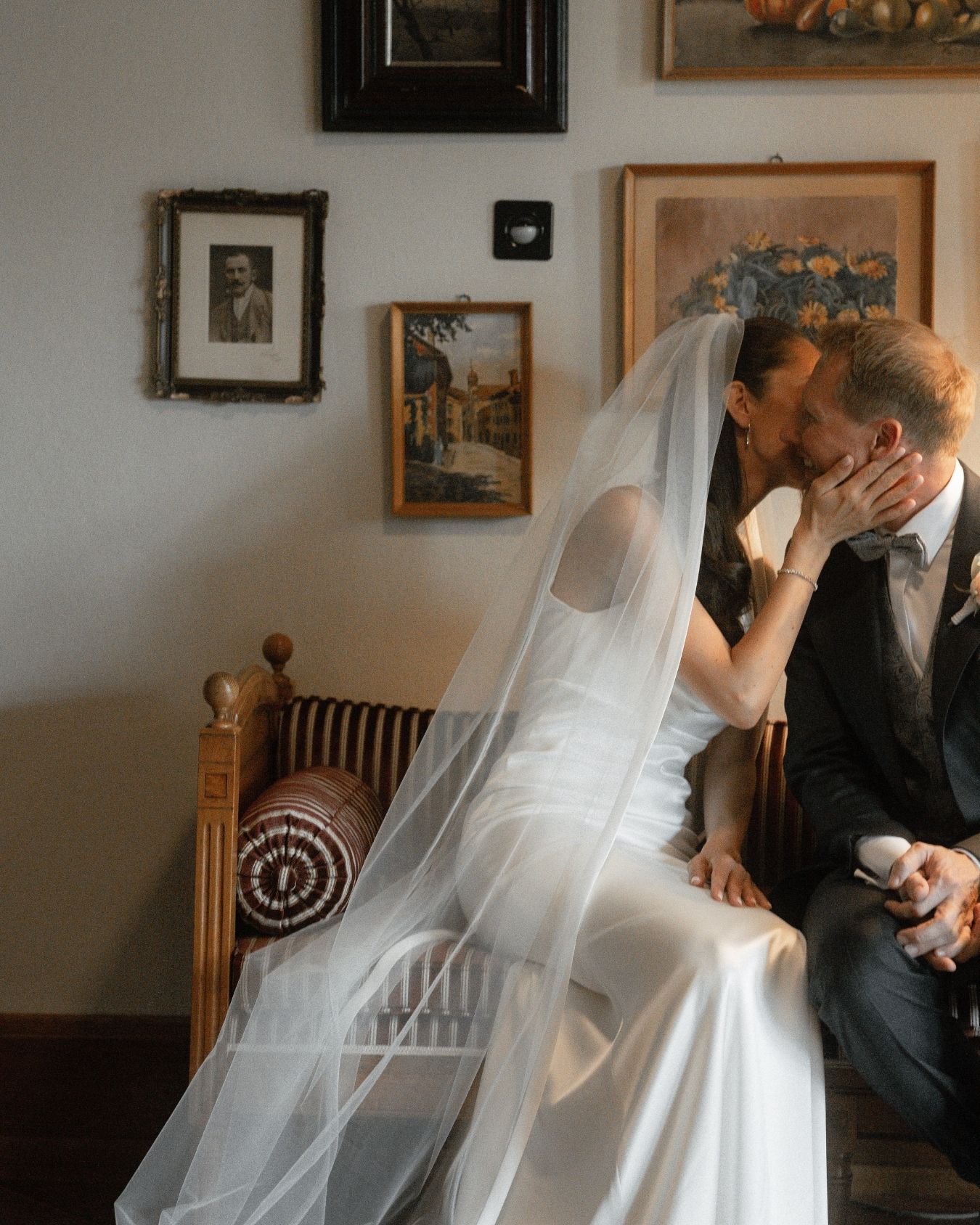 A bride and groom share a romantic kiss while seated on a bench, surrounded by vintage framed artwork on the wall.