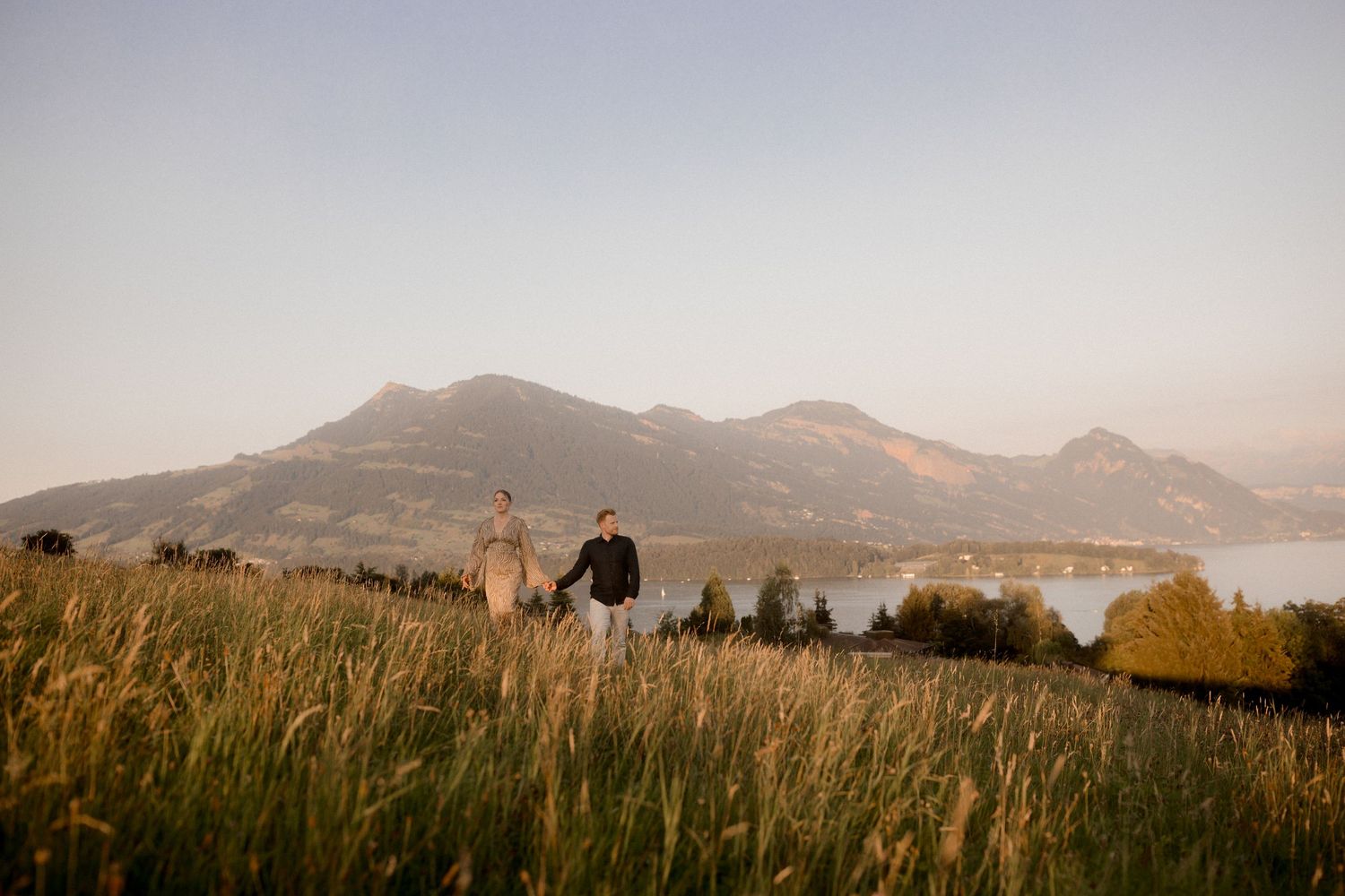 Silhouettes walk through golden grass at sunset with majestic mountain peaks and lake in the background.