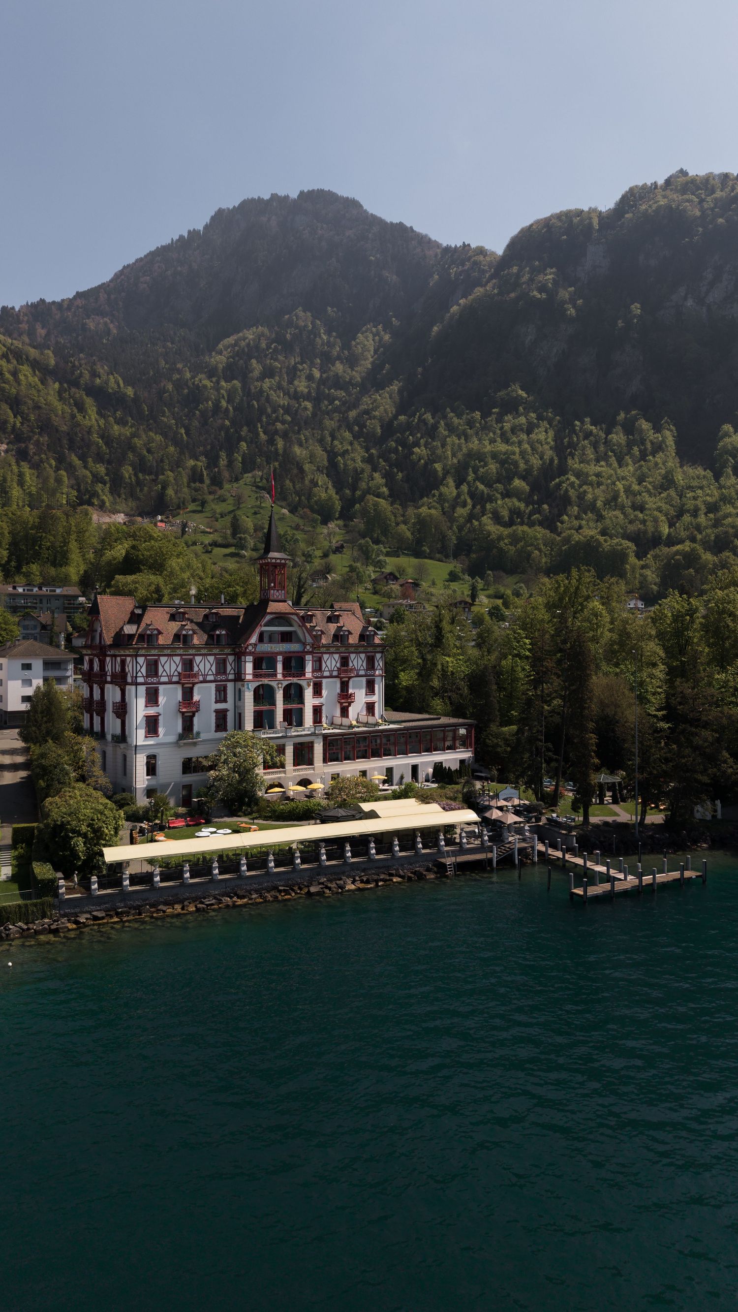 Grand historic hotel on the shores of a Swiss alpine lake surrounded by lush green mountains on a clear sunny day.