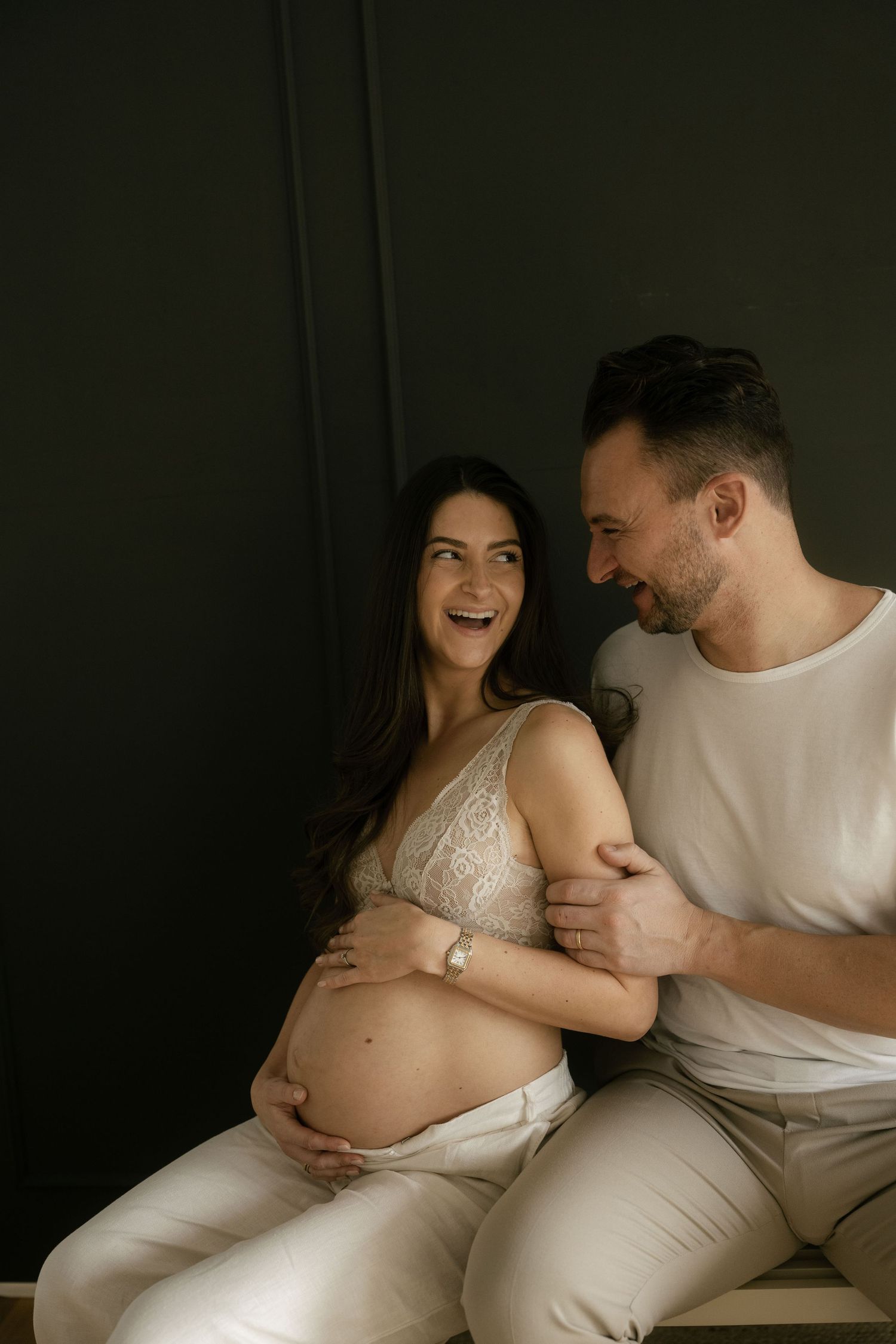 Moody maternity portraits of couple sharing joyful moments against dark background.