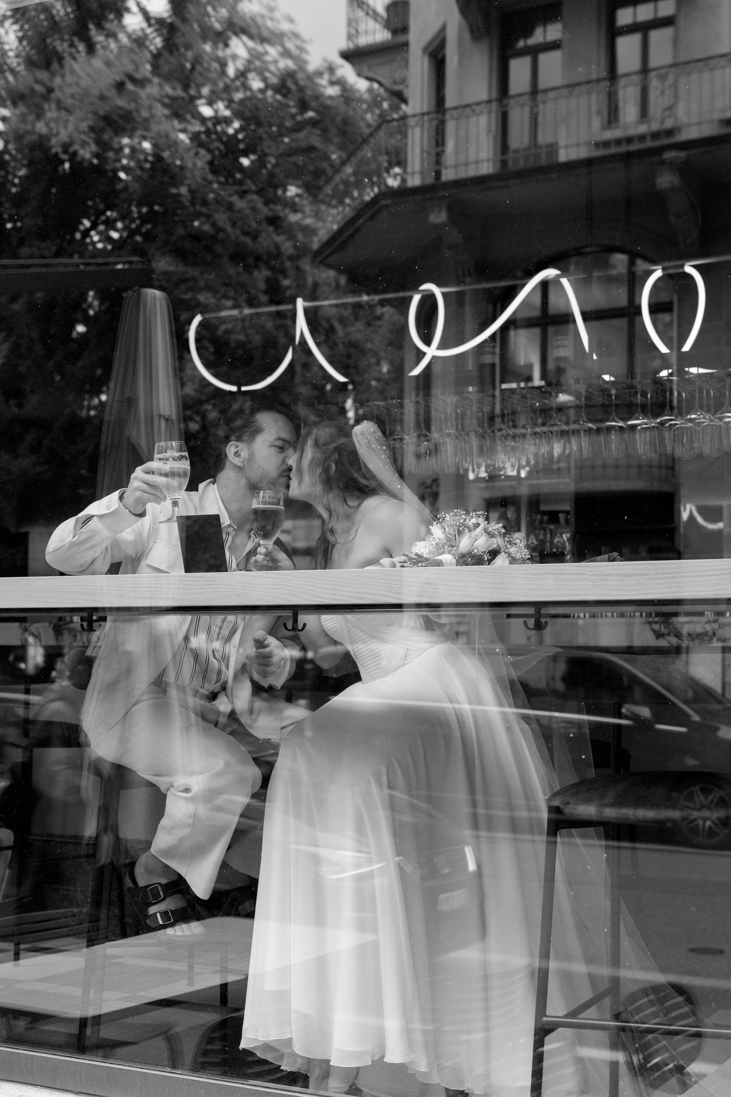 Black and white photo of newlyweds sharing a toast through a cafe window on their wedding day. La bestia Pizza Luzern