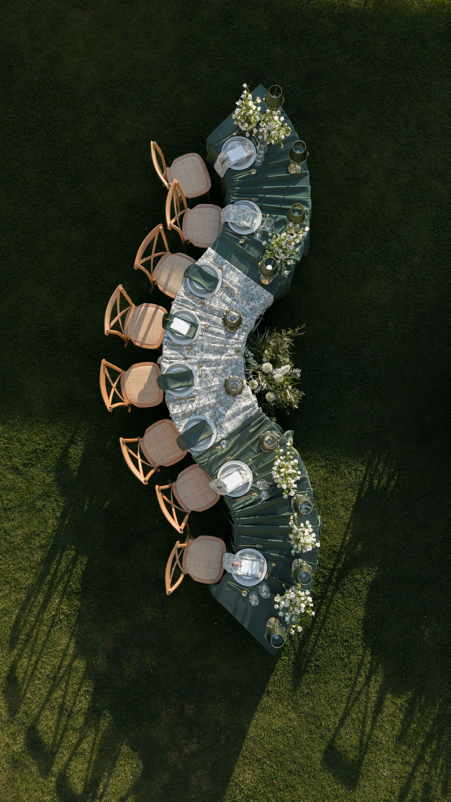 Aerial view of an elegant curved outdoor dining table set with floral arrangements, glassware, and wooden chairs on green grass.