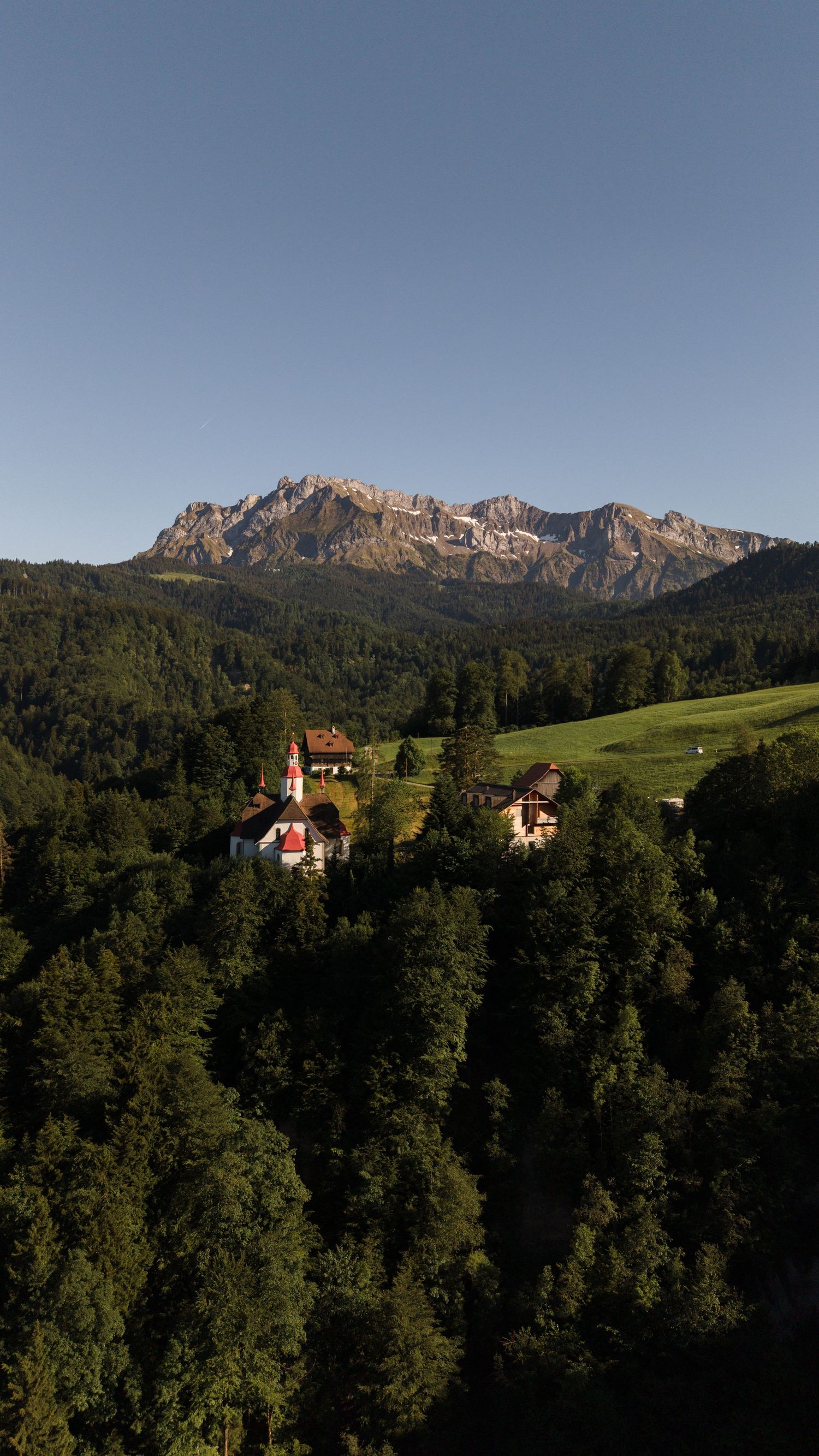 Aerial view of a Alpine village church nestled among lush green forests with dramatic rocky mountain peaks in the background.