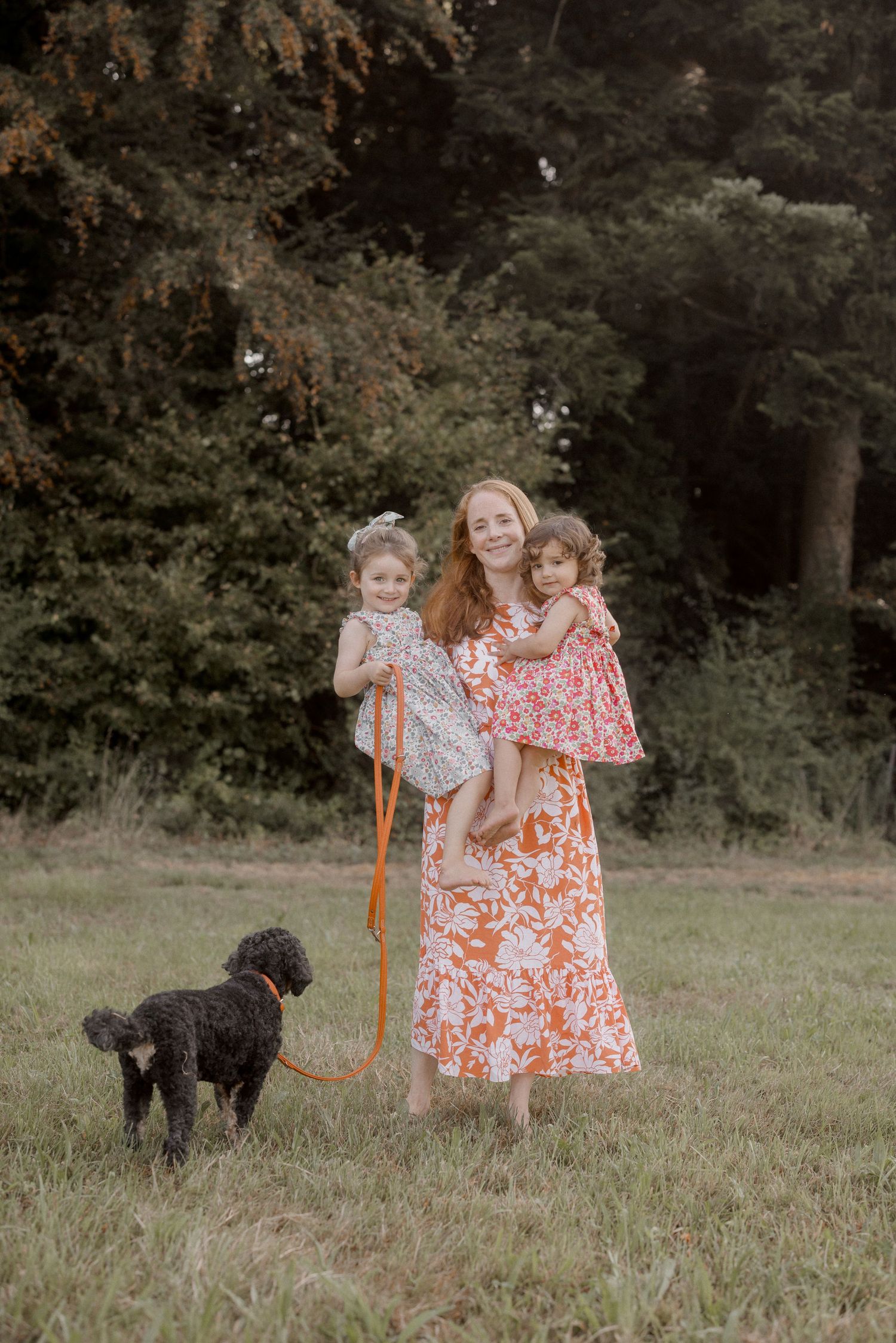 Children in floral dresses exploring a grassy field with a black dog.