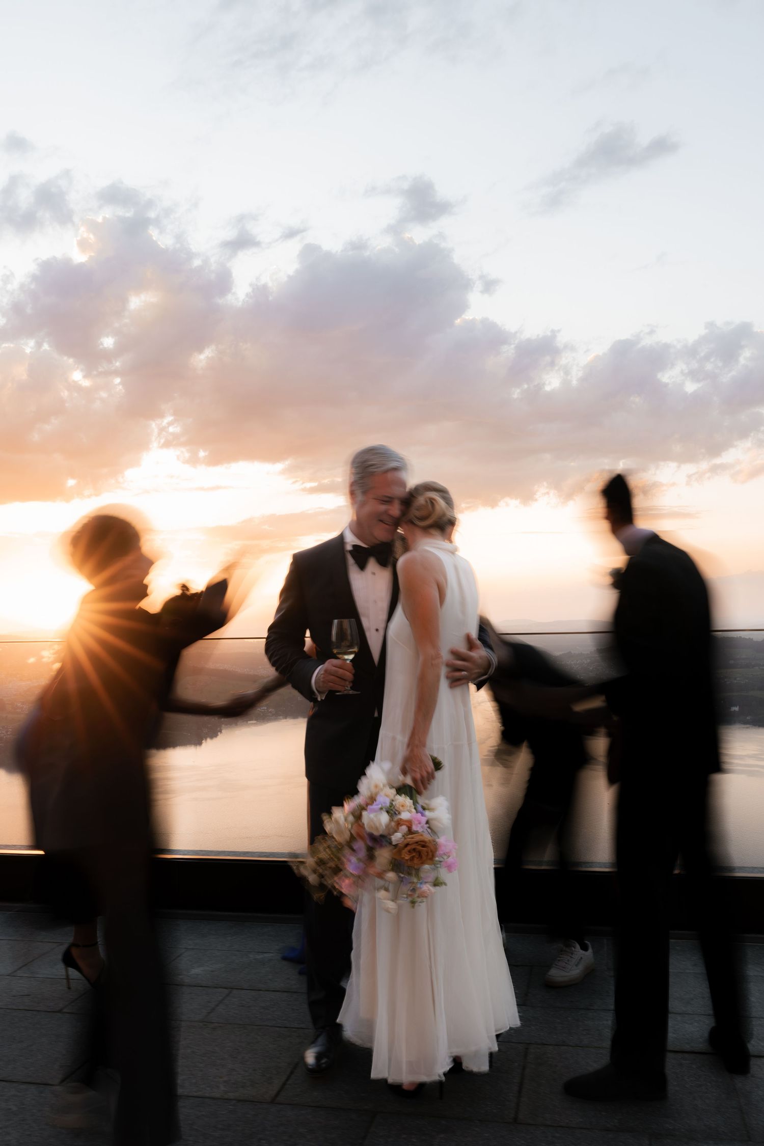 Bride and groom share a romantic moment at sunset on a terrace, surrounded by blurred wedding guests.