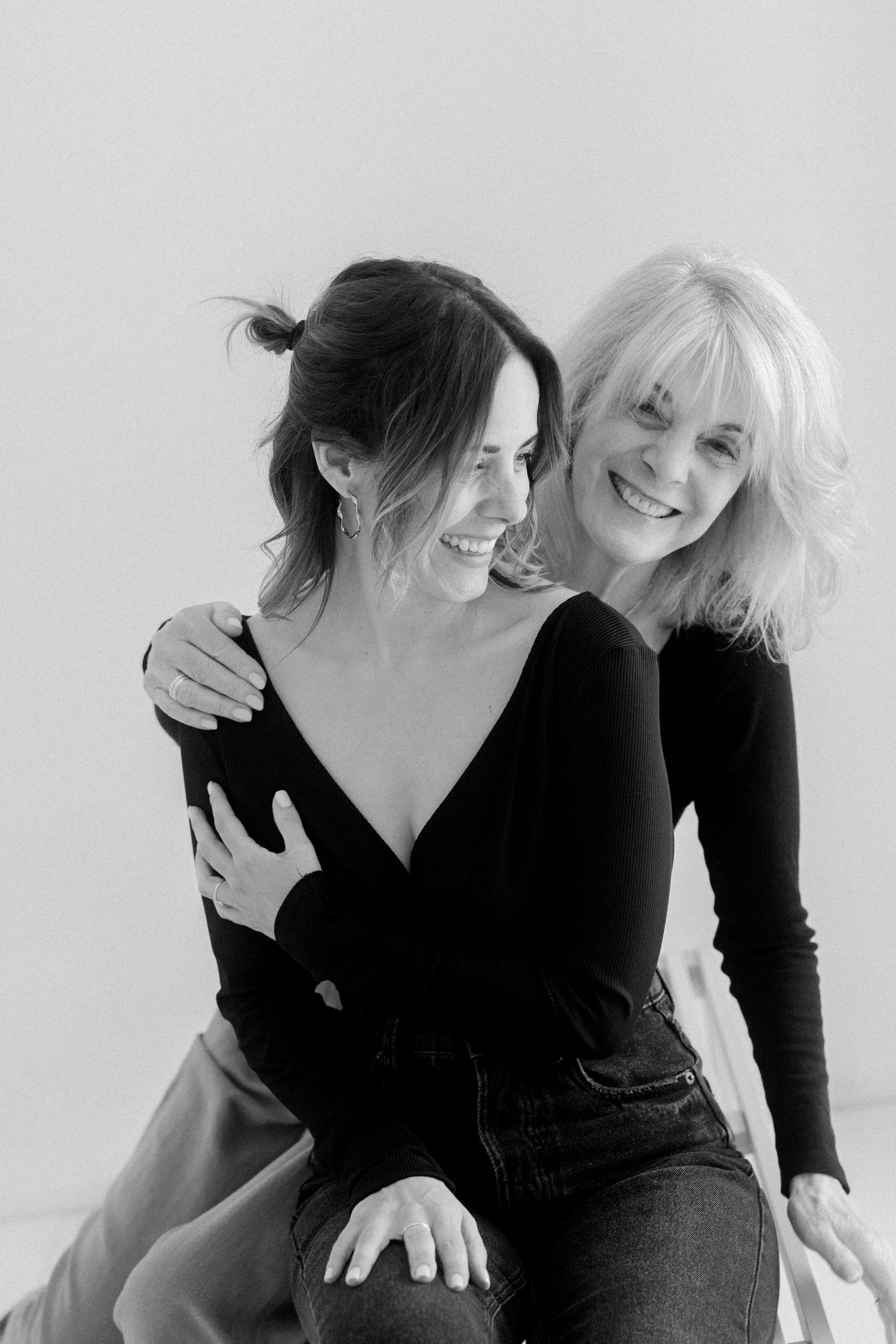 Two people in black clothing share joyful laughter while embracing against a white background.