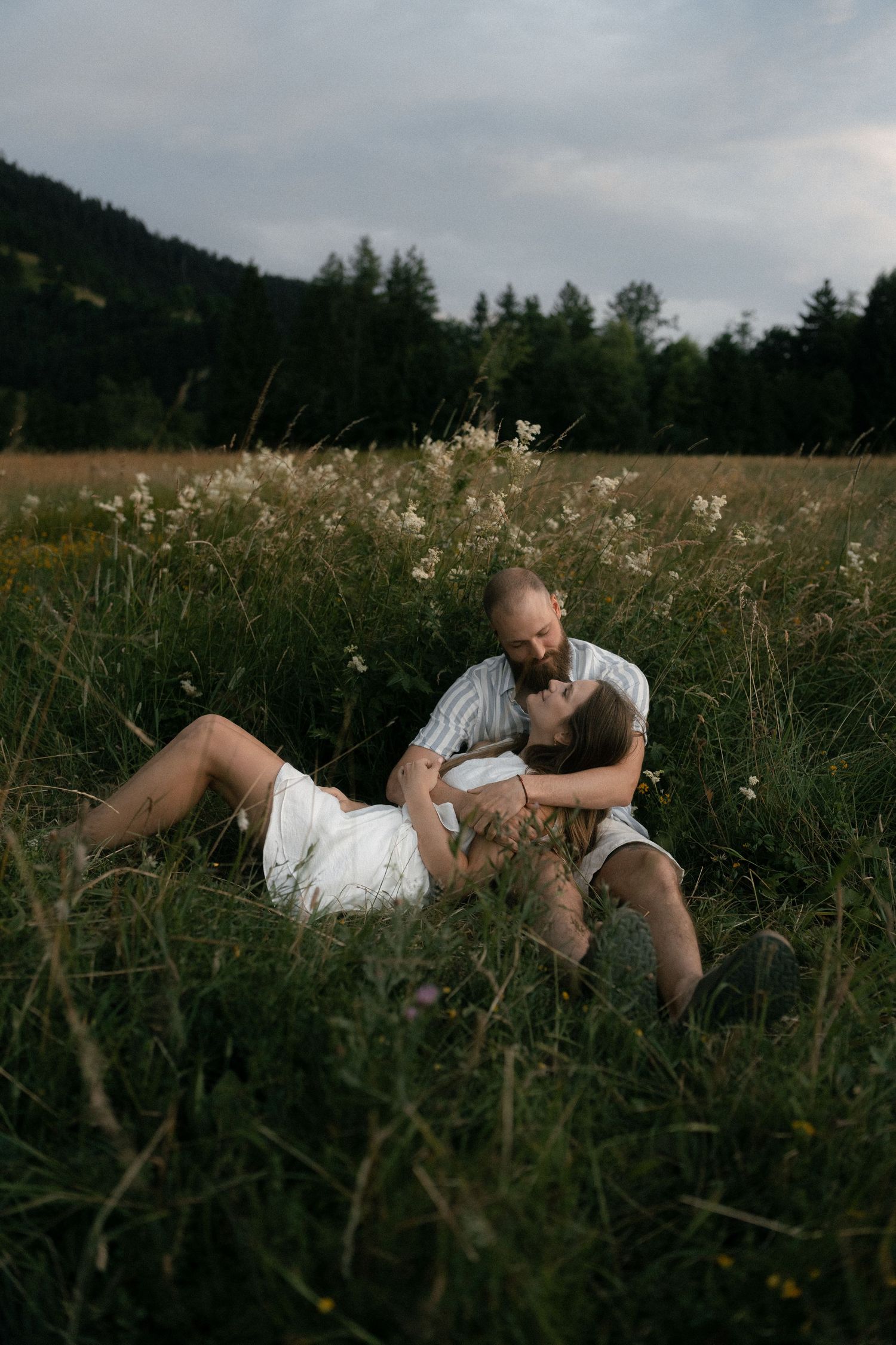 Intimate couple lying in a meadow of tall grass and wildflowers with evergreen trees in the background at dusk.