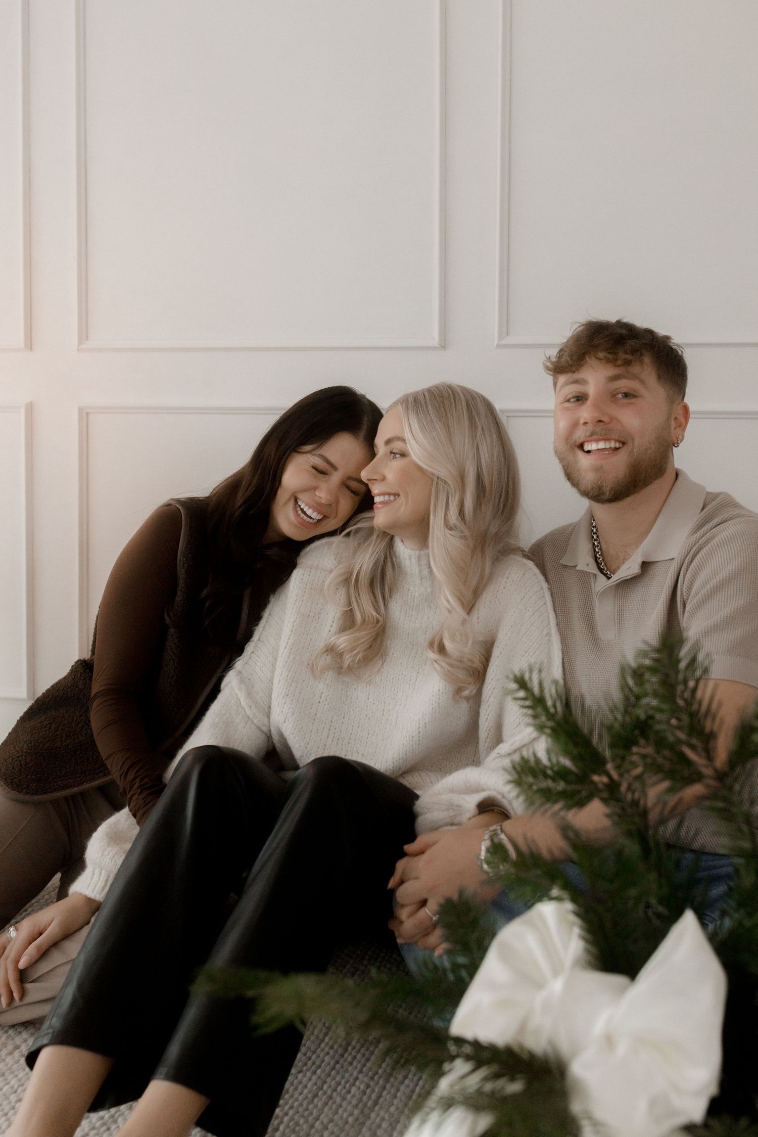 A group of friends laugh together while relaxing on a white couch with a houseplant in the foreground.