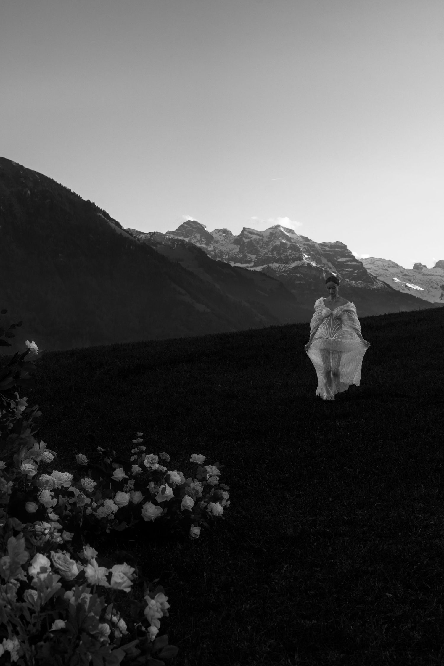 Black and white photo of a bride in white dress standing in mountain meadow with wildflowers at dusk.