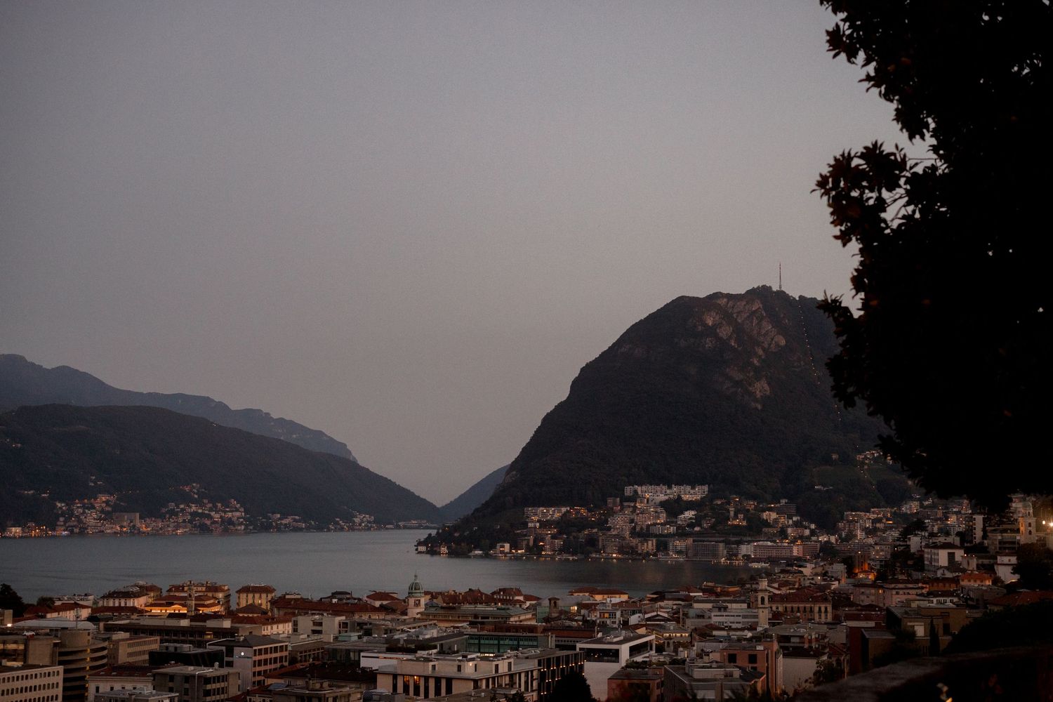 Panoramic dusk view of Lugano, Switzerland, with lake, mountains, and city buildings in soft evening light.