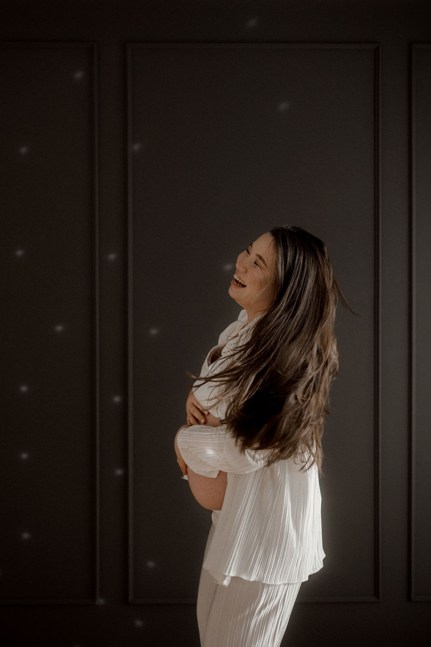 Artistic silhouette portrait of a pregnant woman in white dress against dark background.