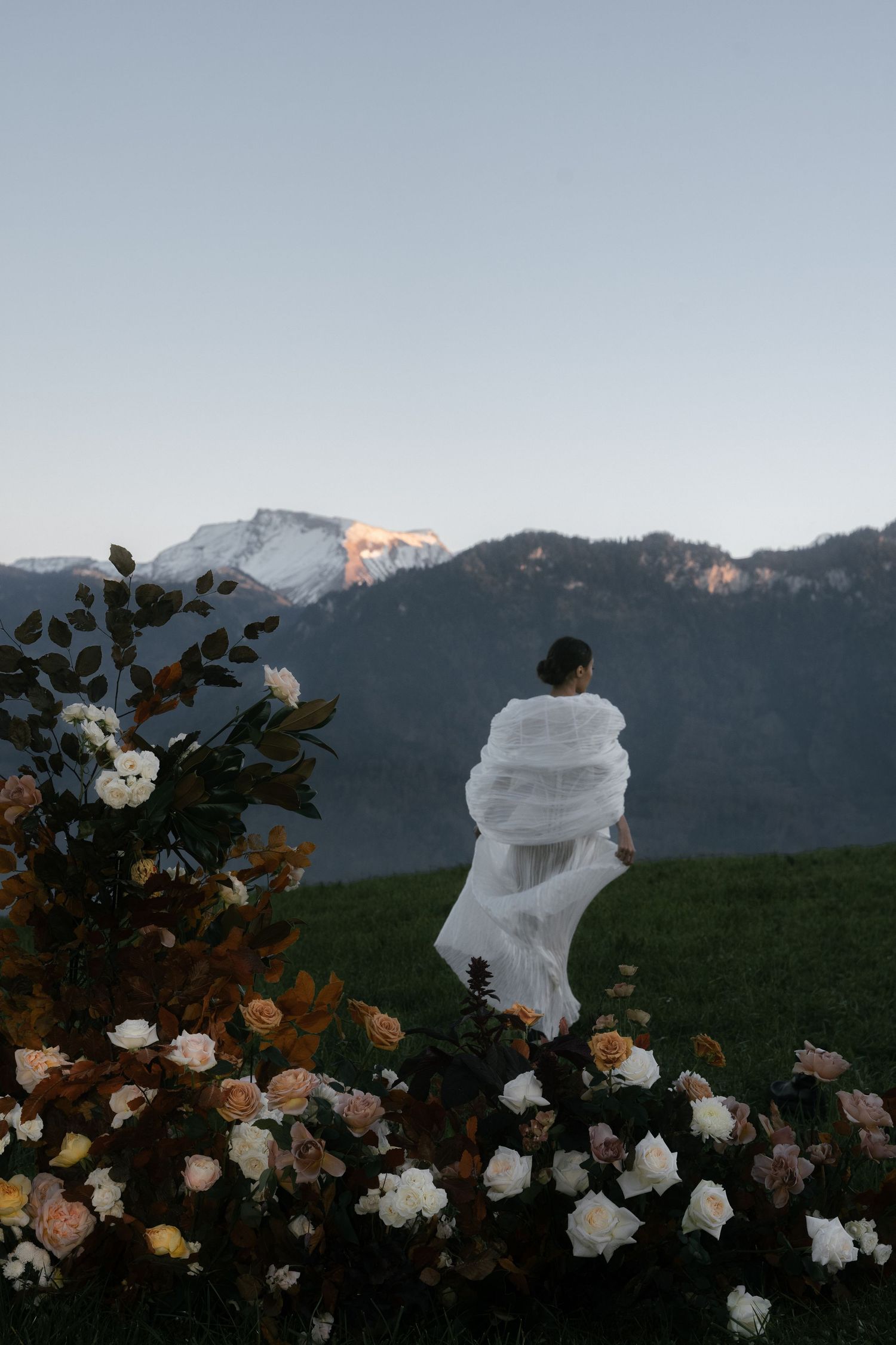 Person in white flowing dress standing in field of white roses with mountain landscape at sunset.
