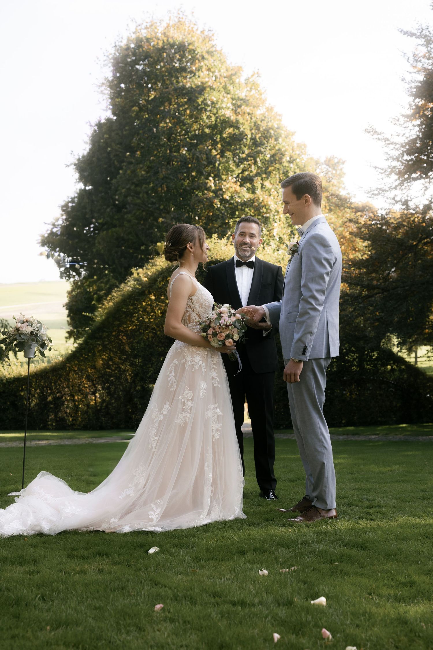 A wedding couple and officiant stand together on a sunlit lawn during an outdoor ceremony at golden hour.
