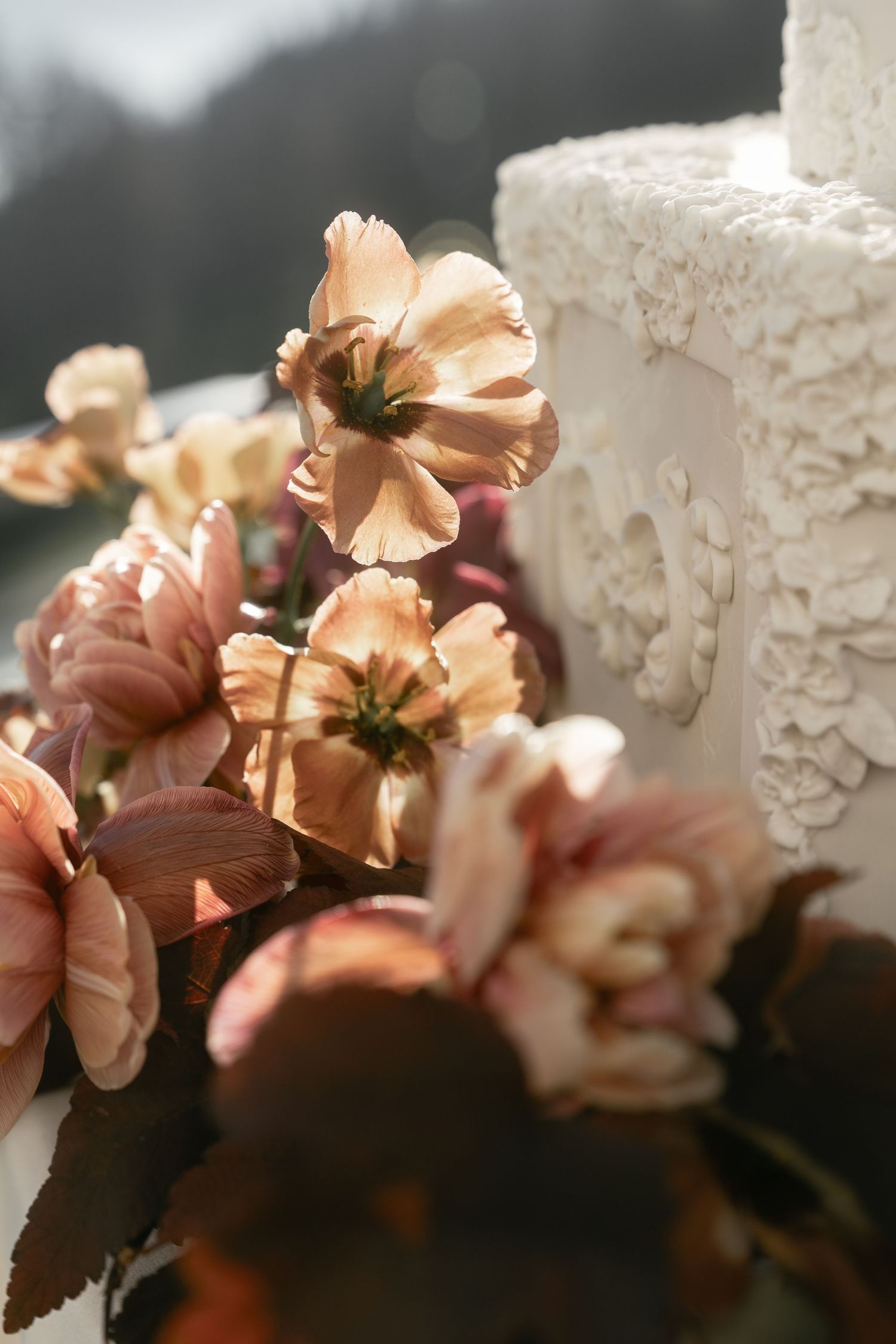 Soft pink flowers beside ornate white stone cross in sunlight.